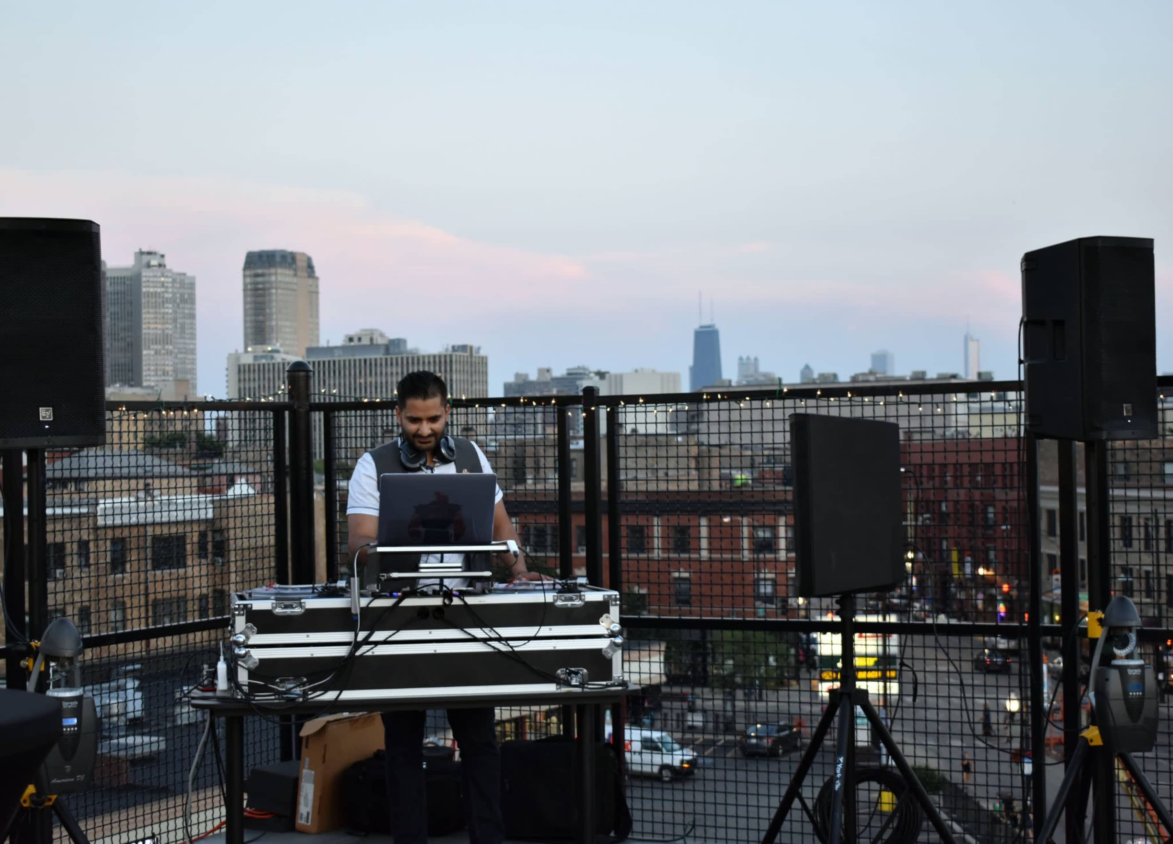 A DJ performs at a rooftop venue with a city skyline in the background during twilight.