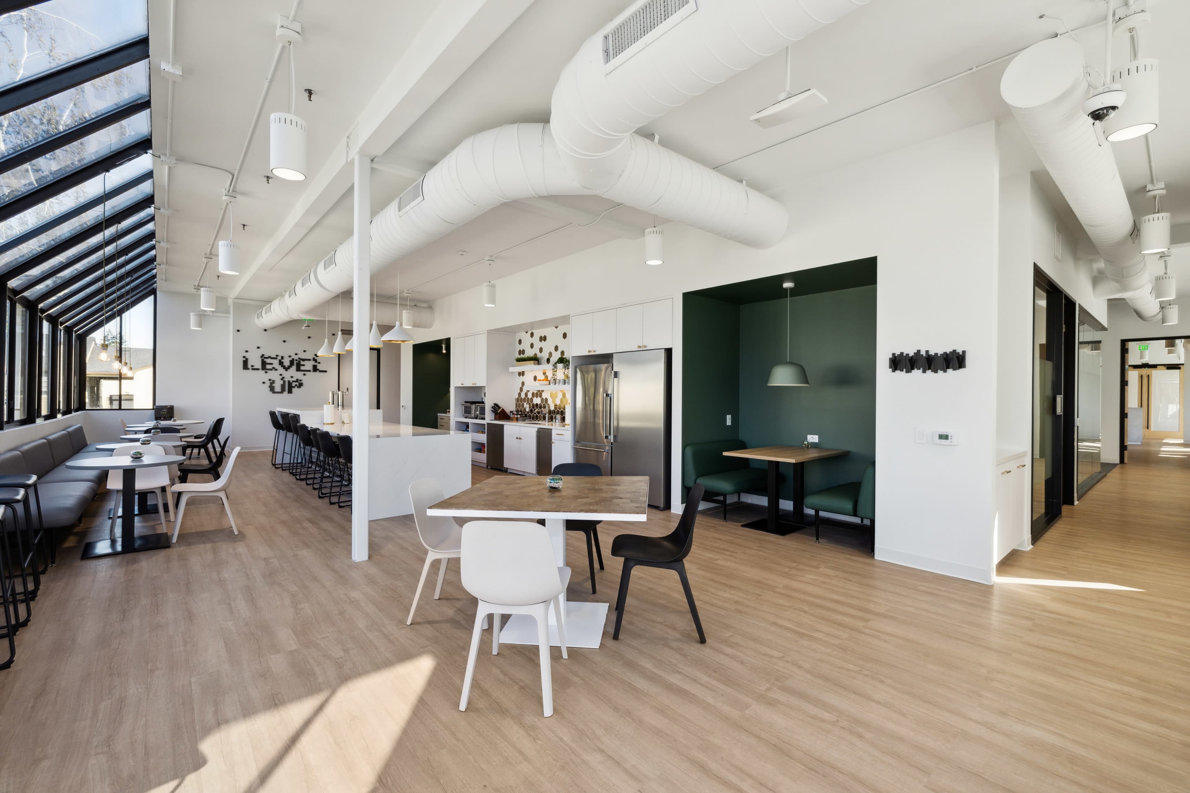 The image shows a modern, well-lit open workspace with a kitchen area, seating arrangements, and large windows.