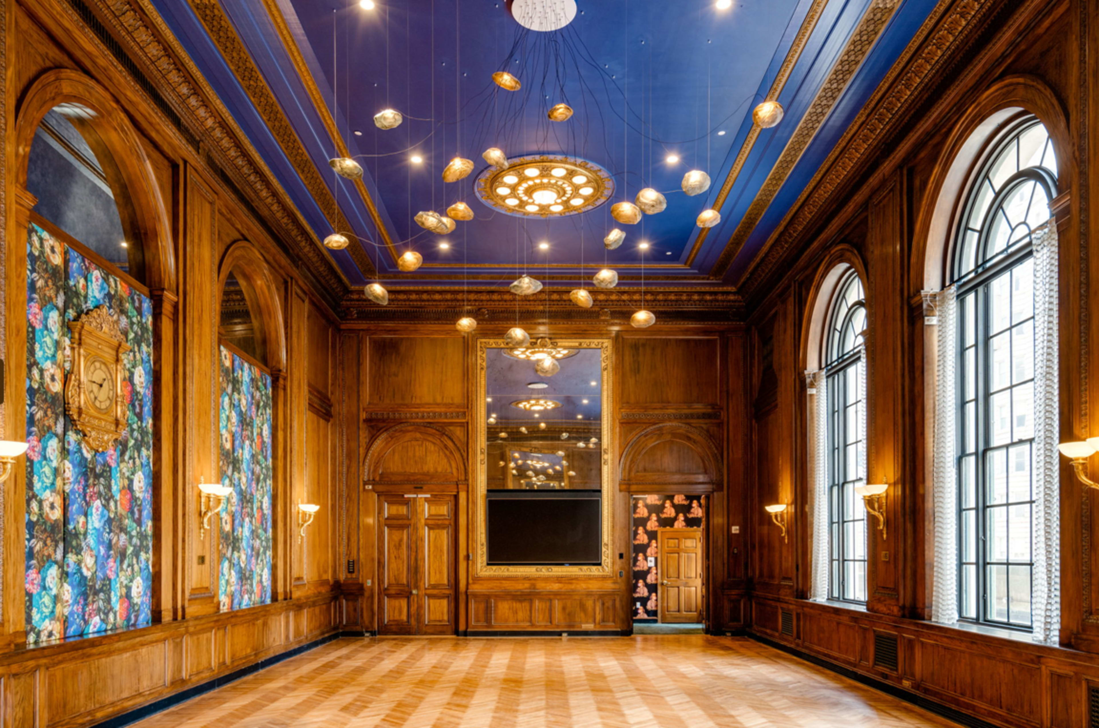 A spacious room with wooden paneling, large windows, and ornate lighting fixtures, featuring a floral wallpaper on one wall.