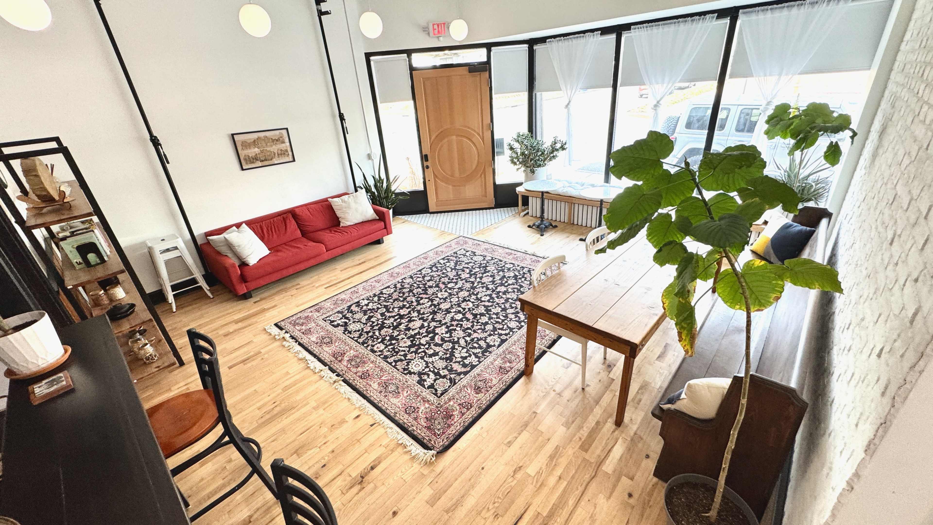 The image depicts a bright interior space with a red sofa, a large area rug, a wooden table, plants, and large windows allowing natural light.