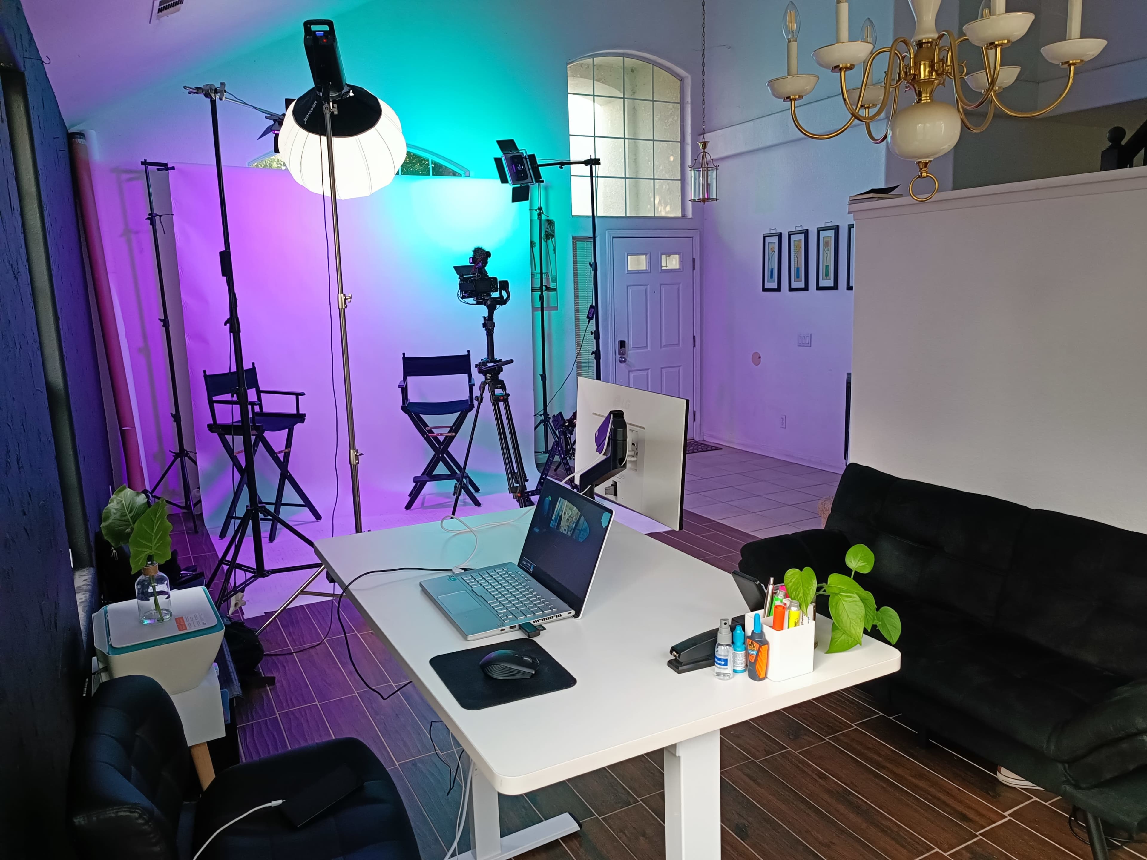 A well-lit studio space featuring a desk with a laptop, colorful backdrop lighting, camera equipment, and two director's chairs.