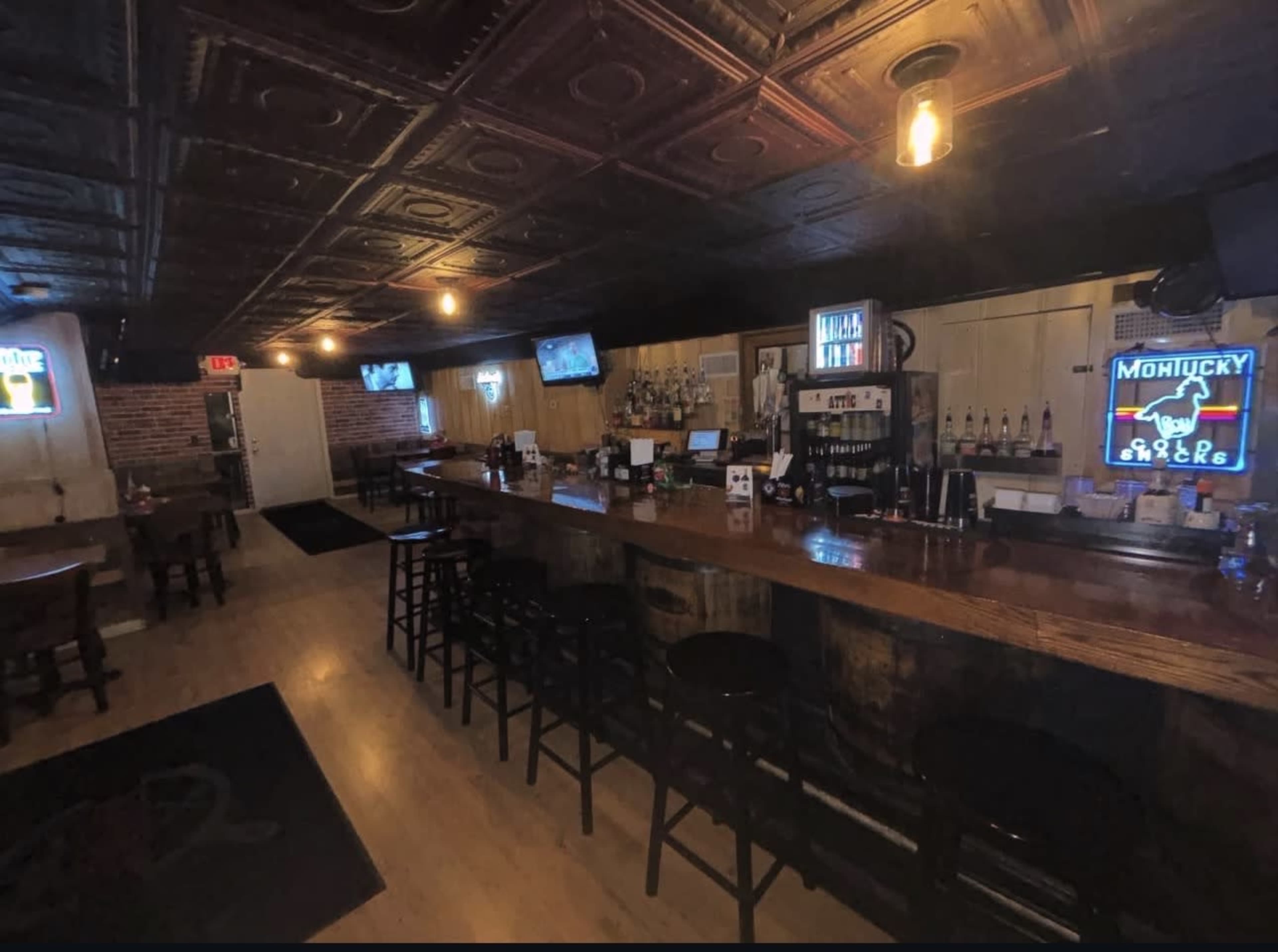 The image shows the interior of a bar with a wooden counter, several stools, and illuminated signs, along with a brick wall and multiple televisions mounted on the walls.