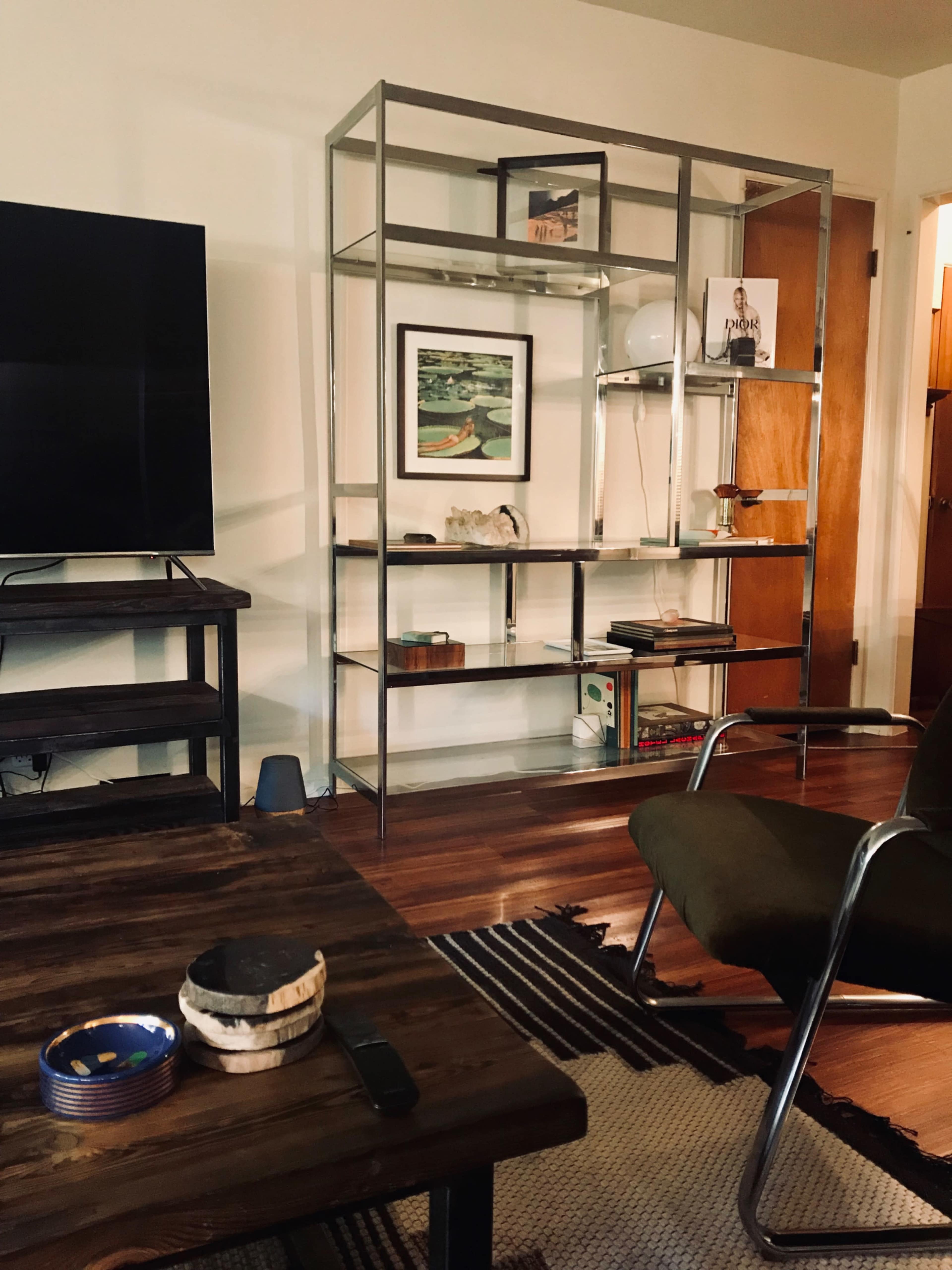 The image shows a living room with a metallic shelving unit displaying various decorative items, a wooden coffee table with a small bowl, and a green armchair.
