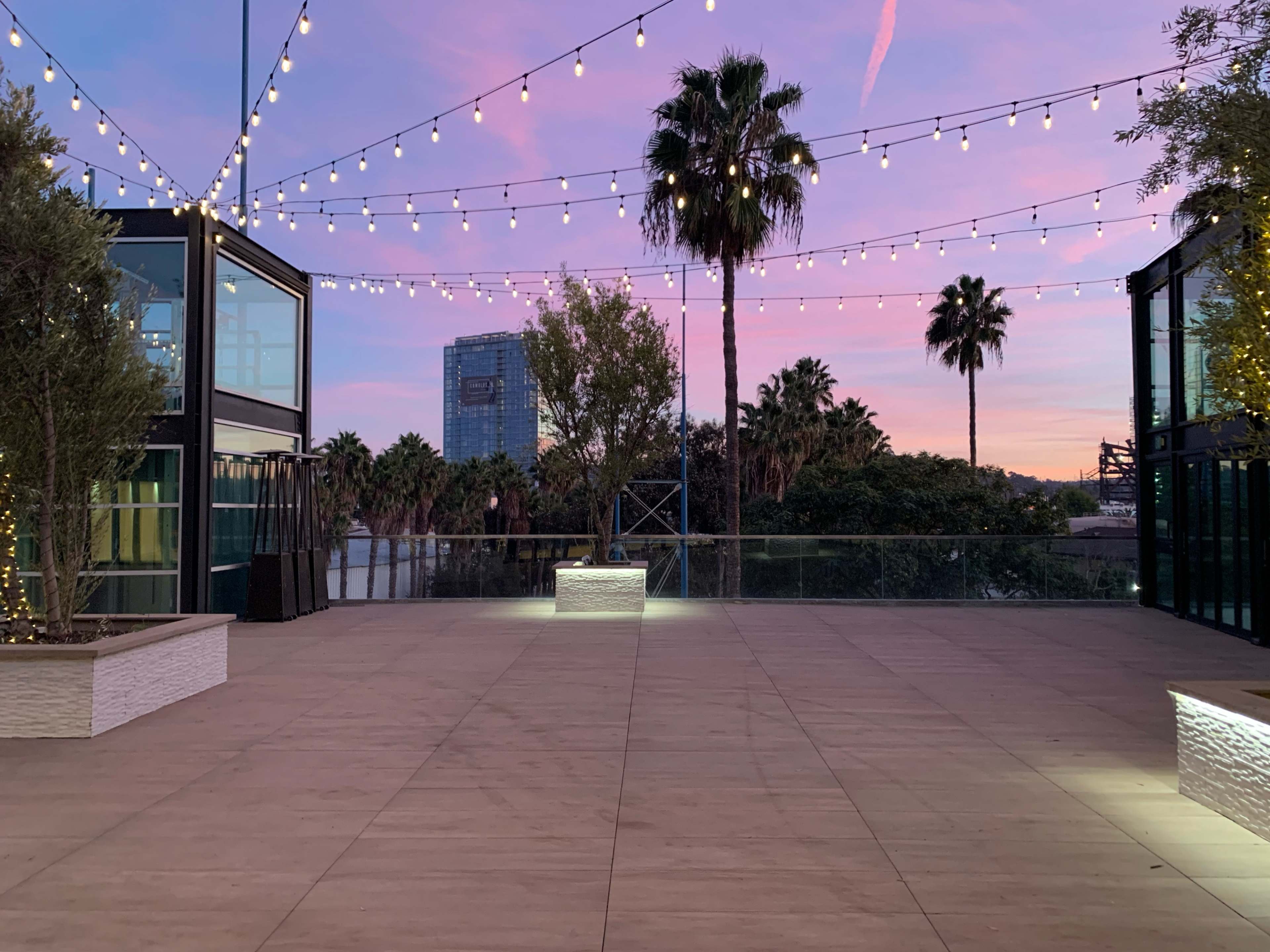 A spacious outdoor area with string lights overhead, palm trees in the background, and a colorful sunset sky.