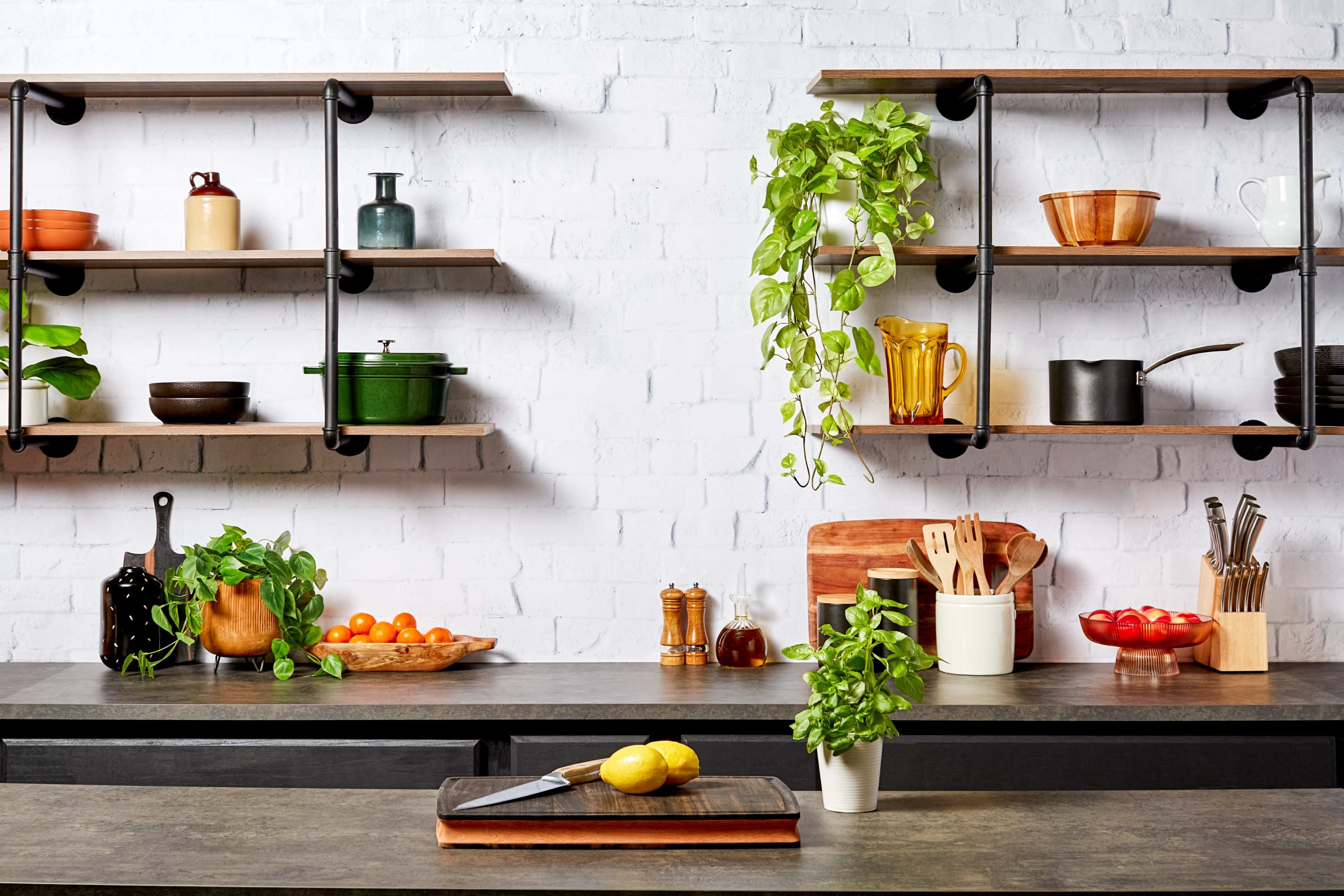 A well-organized kitchen features open shelves with various dishes and plants, a cutting board with lemons, and bowls of fruits on the counter.