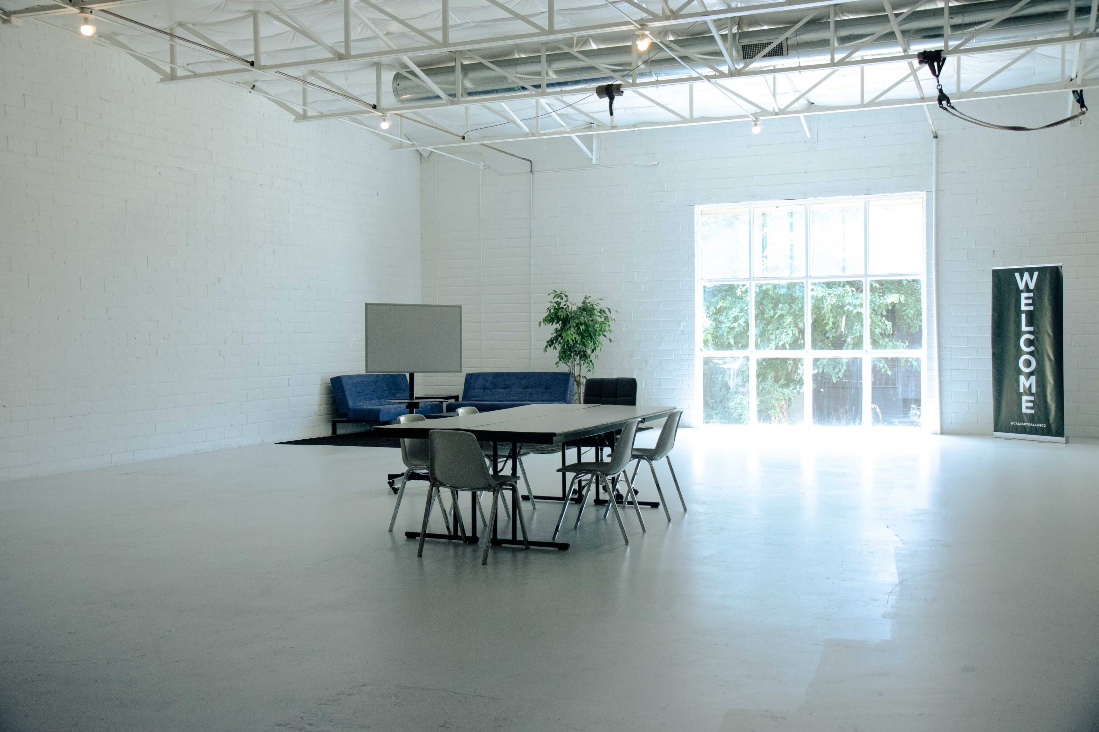 A spacious, minimalist room with a large table surrounded by chairs, a sofa in the corner, and a large window letting in natural light.