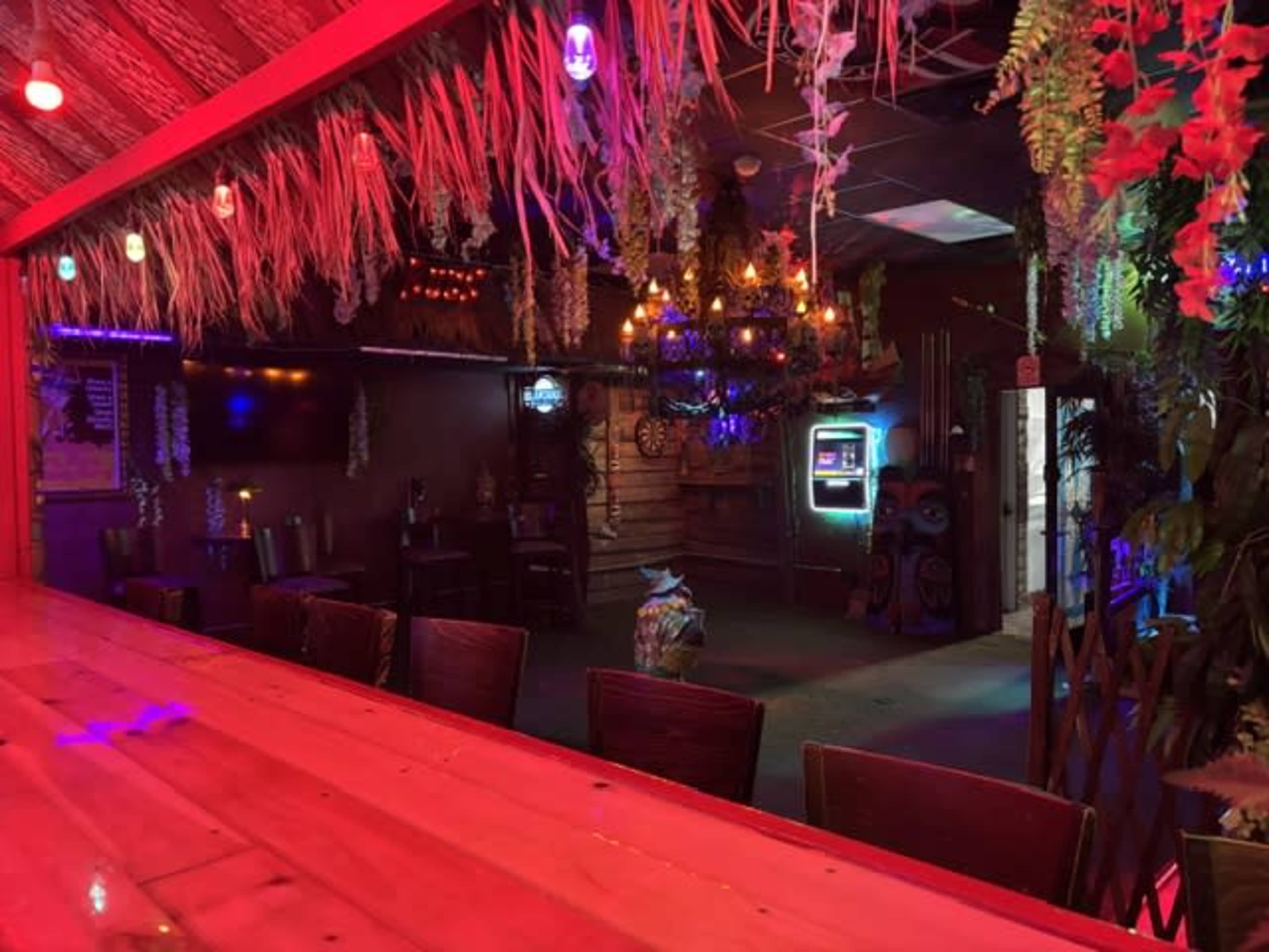 The interior of a dimly lit bar features wooden furniture, colorful LED lights, and tropical decorations.