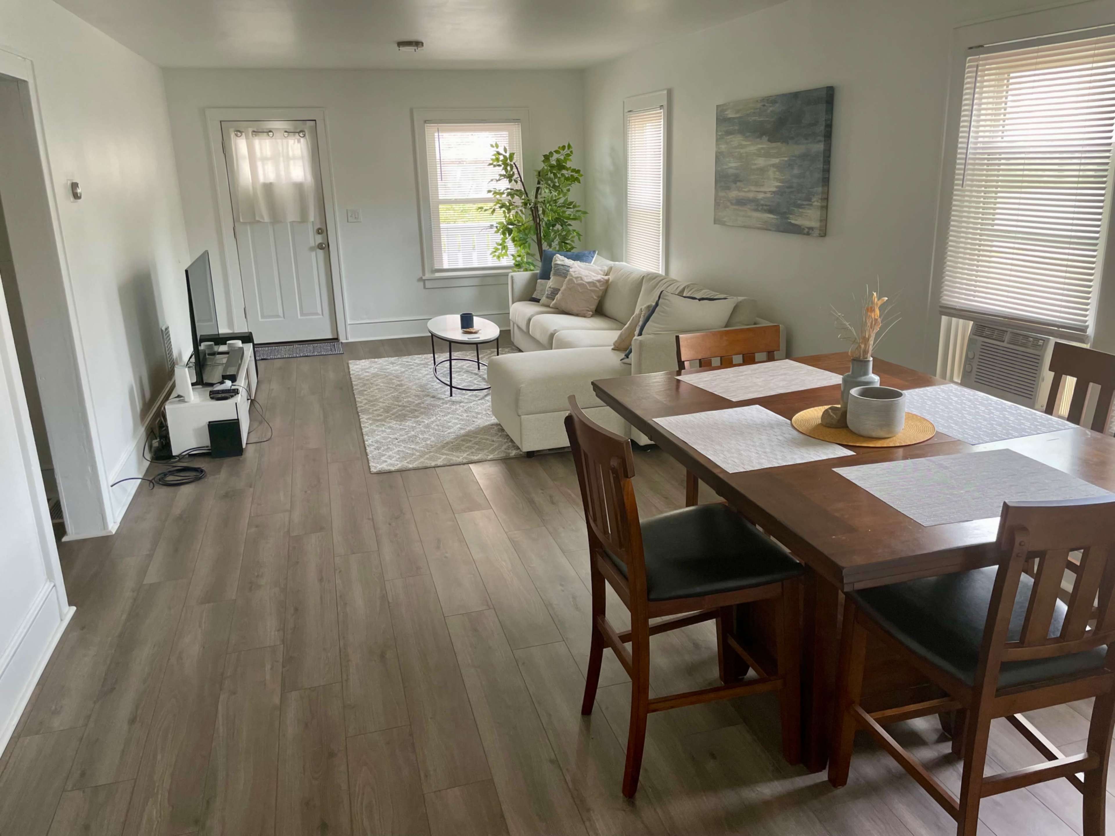 The image shows a bright, minimalist living room and dining area with a sofa, coffee table, dining table, and a television.
