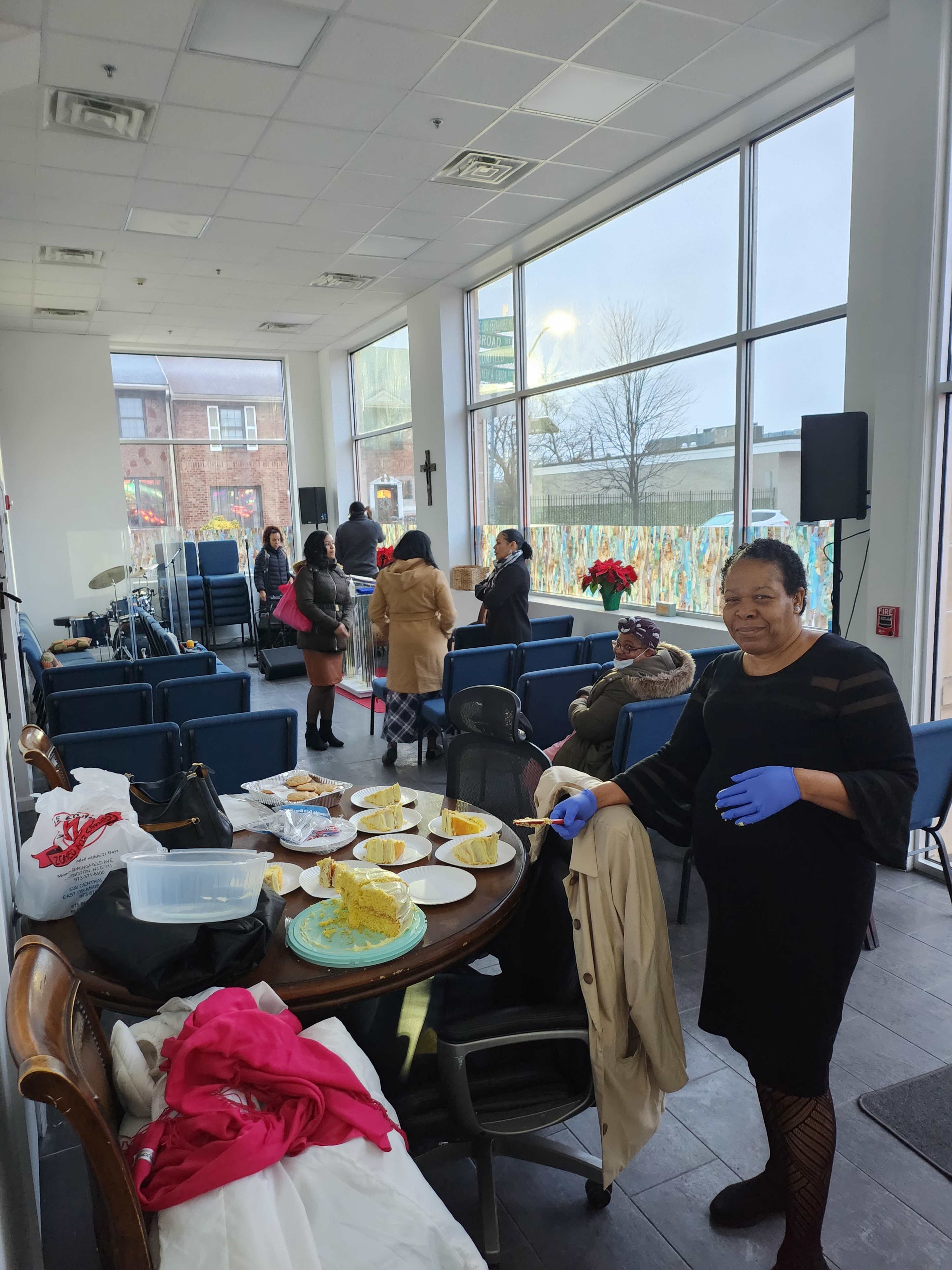 A group of people mingles in a well-lit community space, while a woman wearing gloves serves food at a table.
