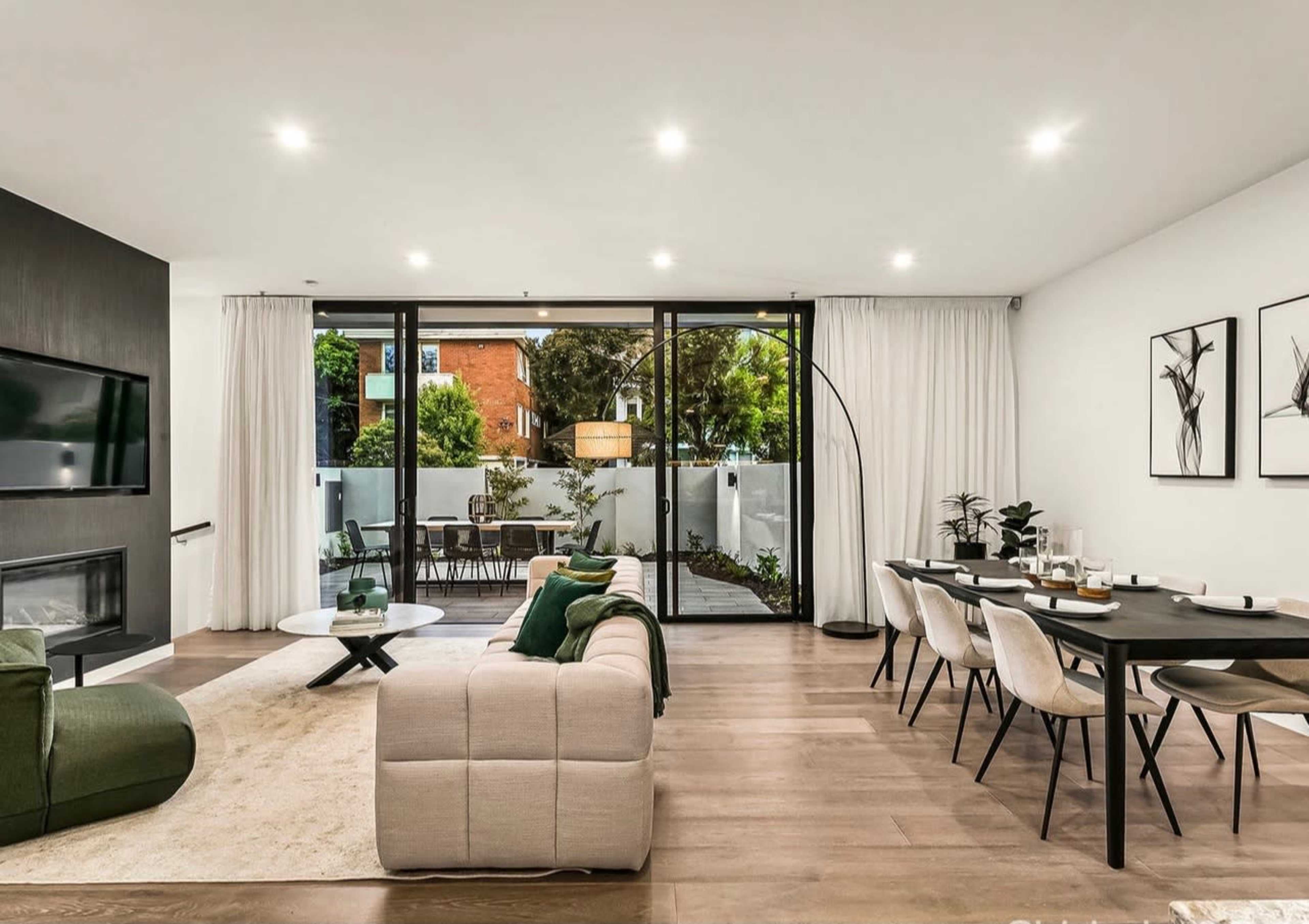 A modern living room features a sectional sofa, a coffee table, a dining area with a black table, and large glass doors opening to an outdoor space with greenery.