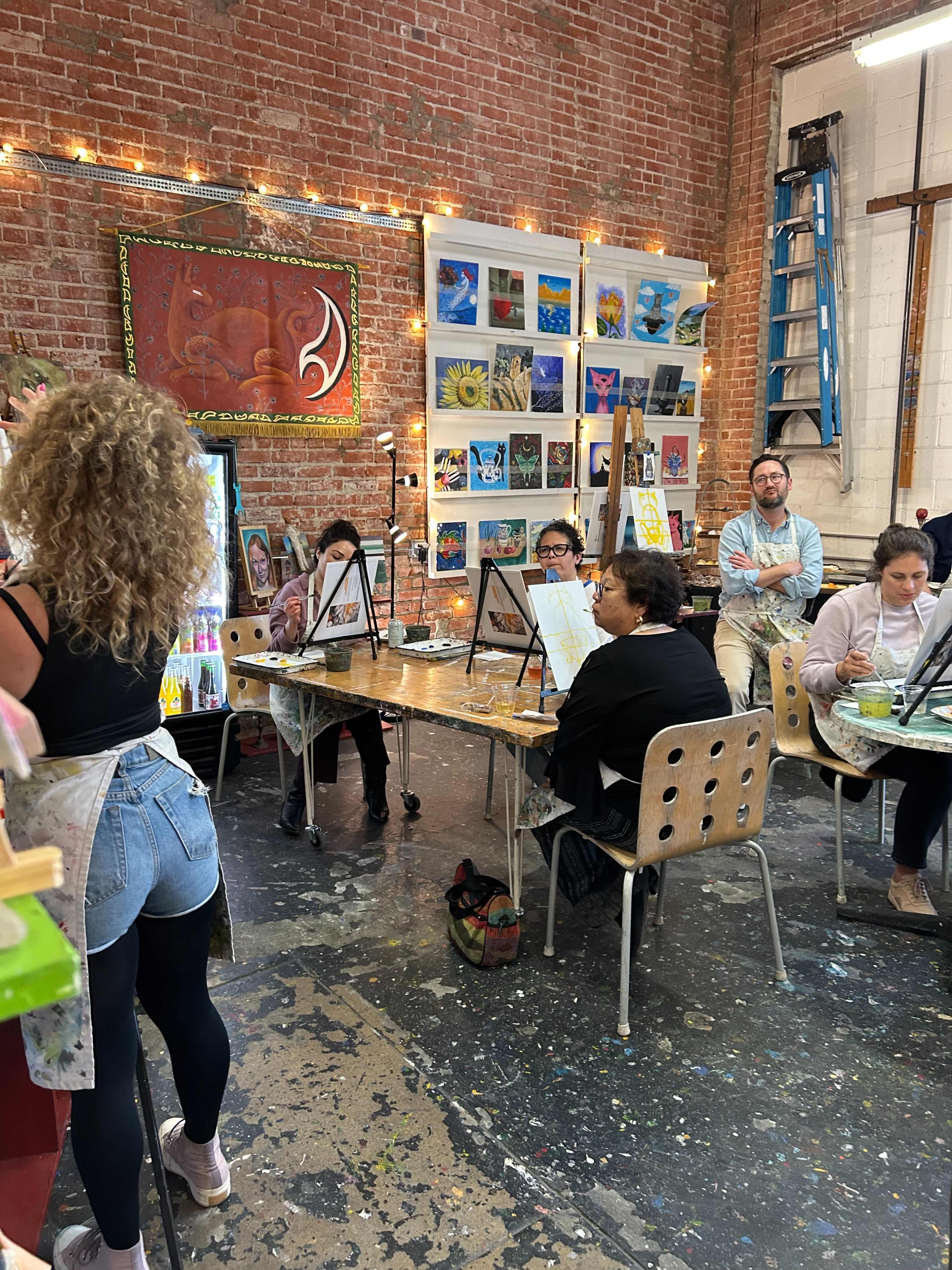 A group of people participates in a painting class inside a studio with exposed brick walls and various artworks displayed.