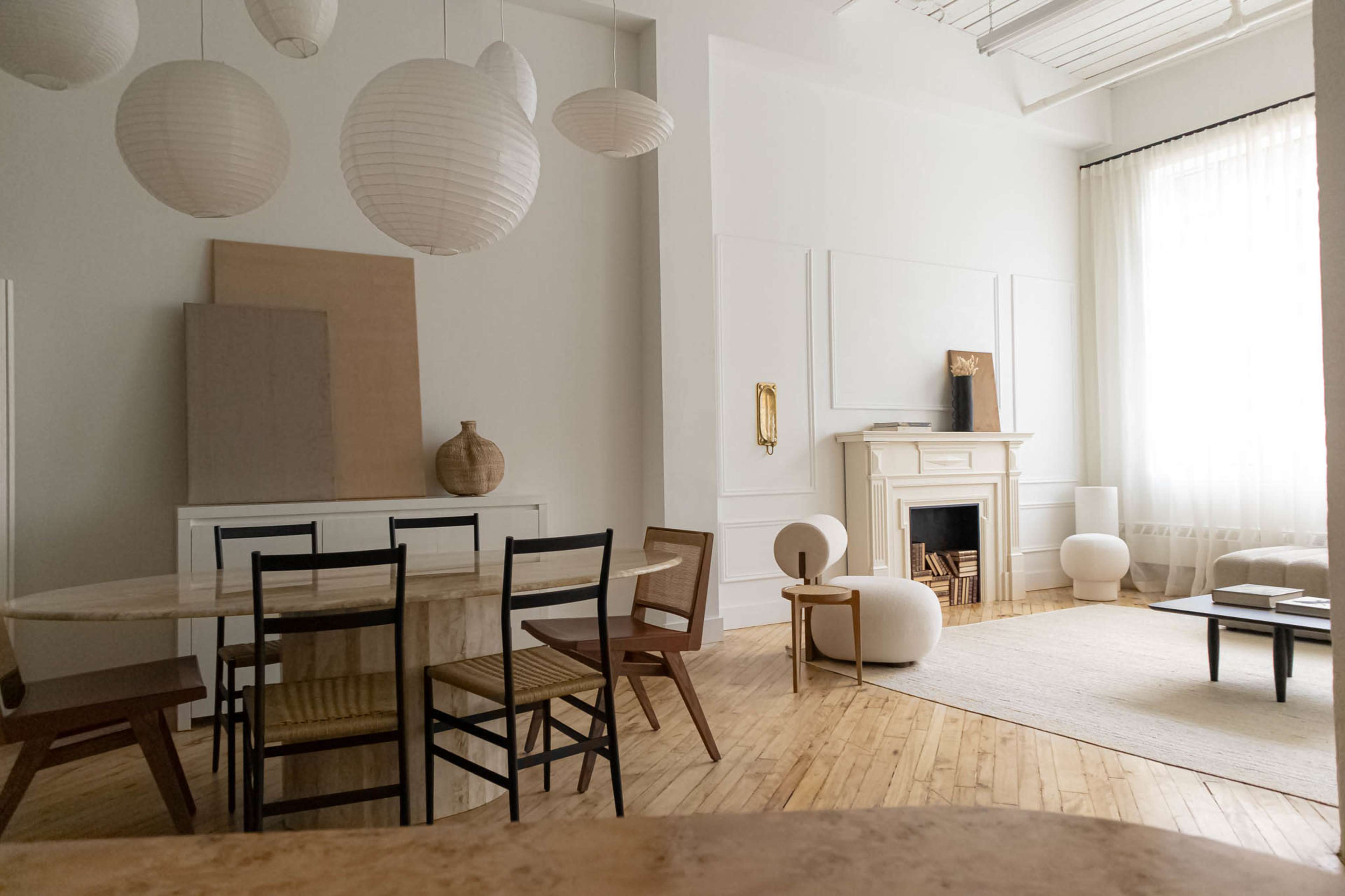 A spacious, minimalist living area features a dining table with chairs, a soft-colored rug, and a fireplace, illuminated by natural light through large windows.
