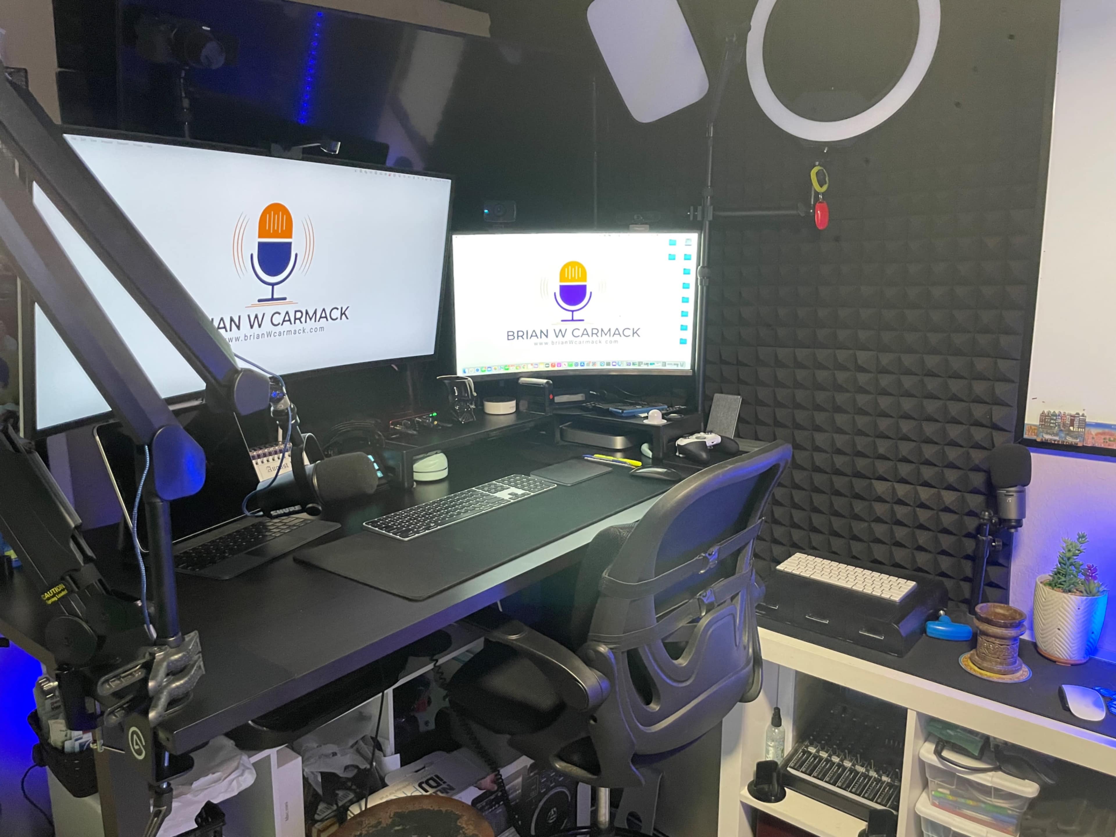 A home office setup featuring two monitors, a microphone, various tech equipment, and soundproofing panels on the walls.