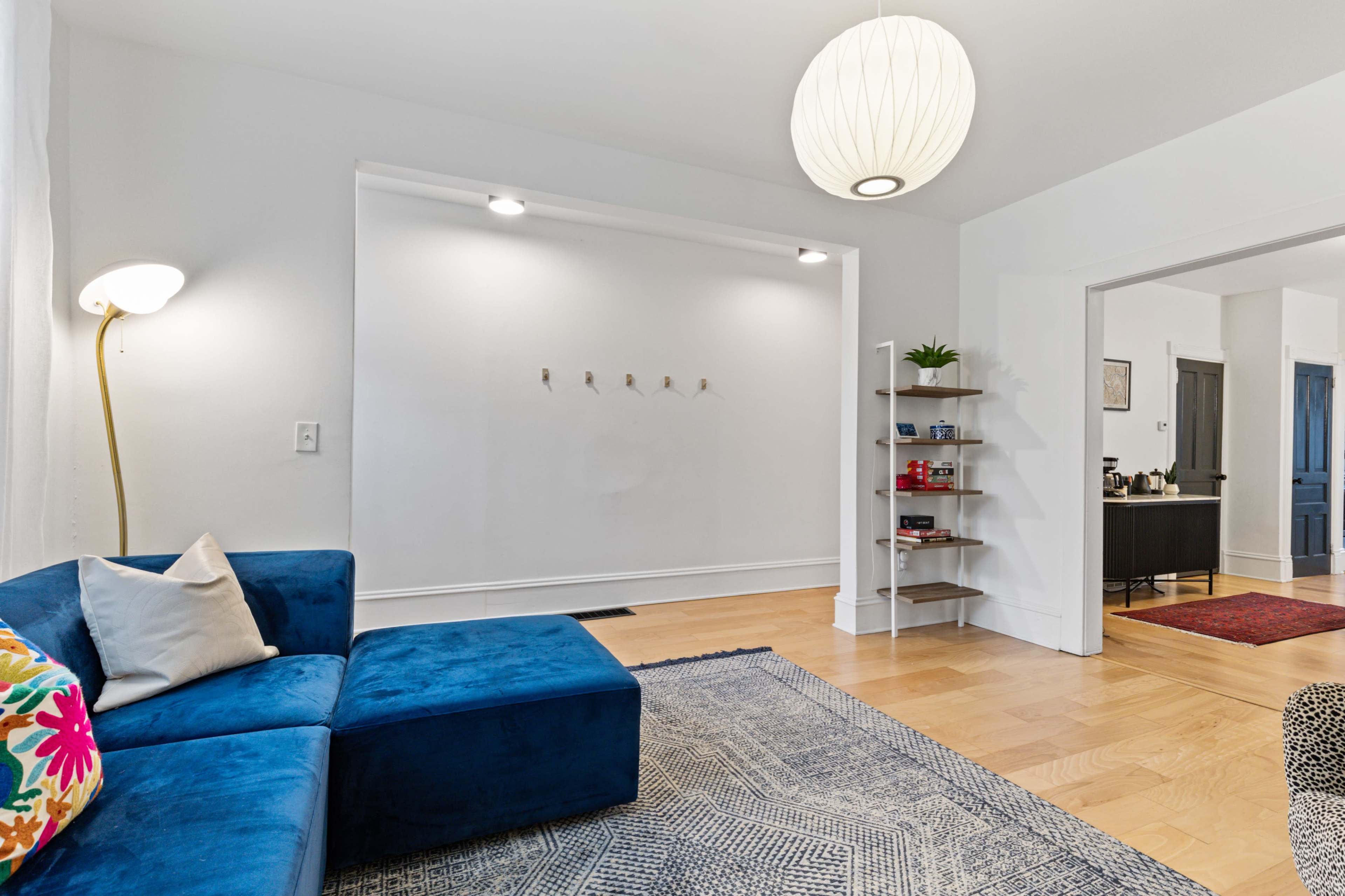 A modern living room features a blue sectional sofa, a floor lamp, and a wall shelf, with a bright pendant light overhead.
