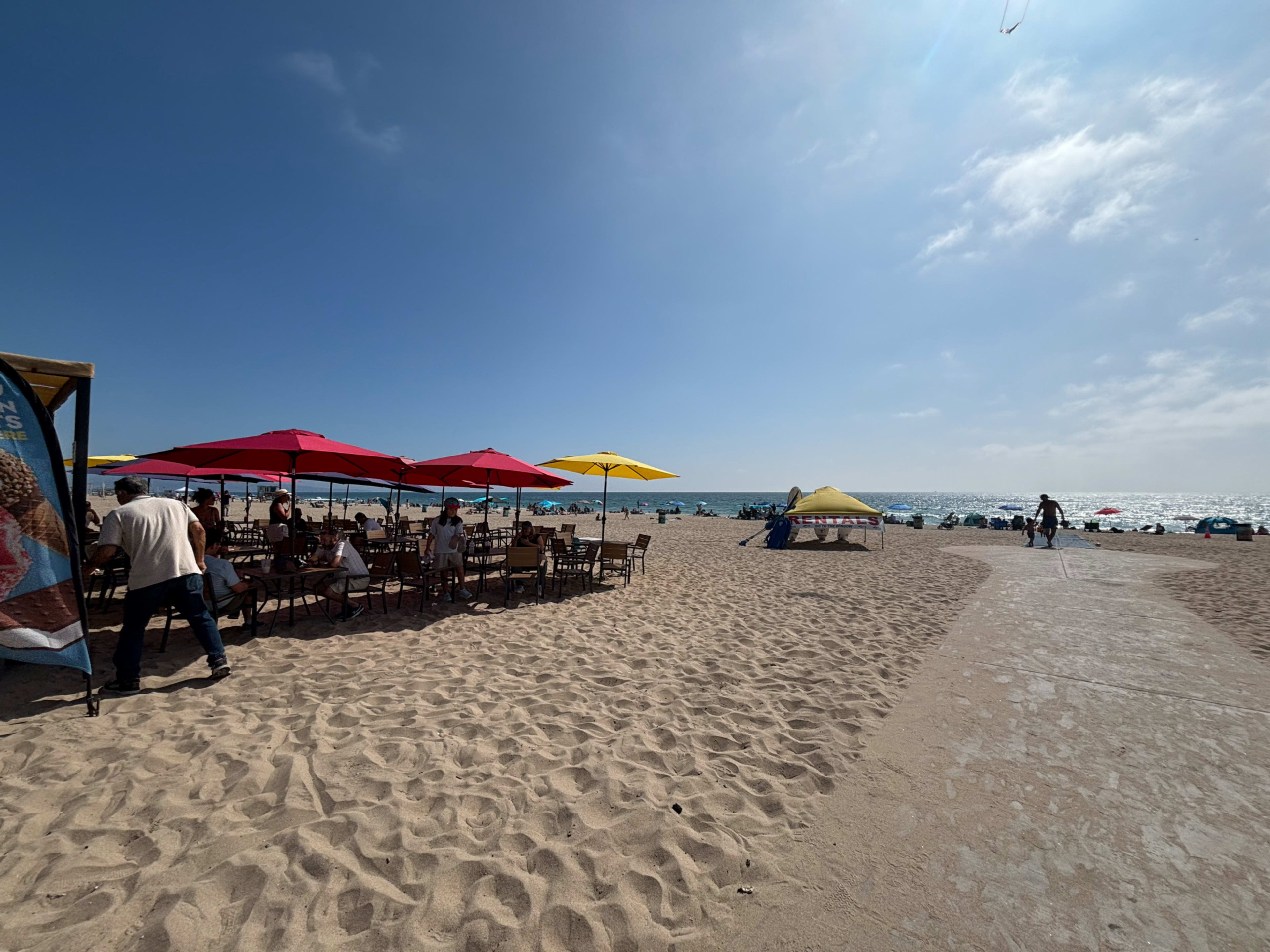 The image shows a sandy beach with people dining under colorful umbrellas and beachgoers near the water in the background.
