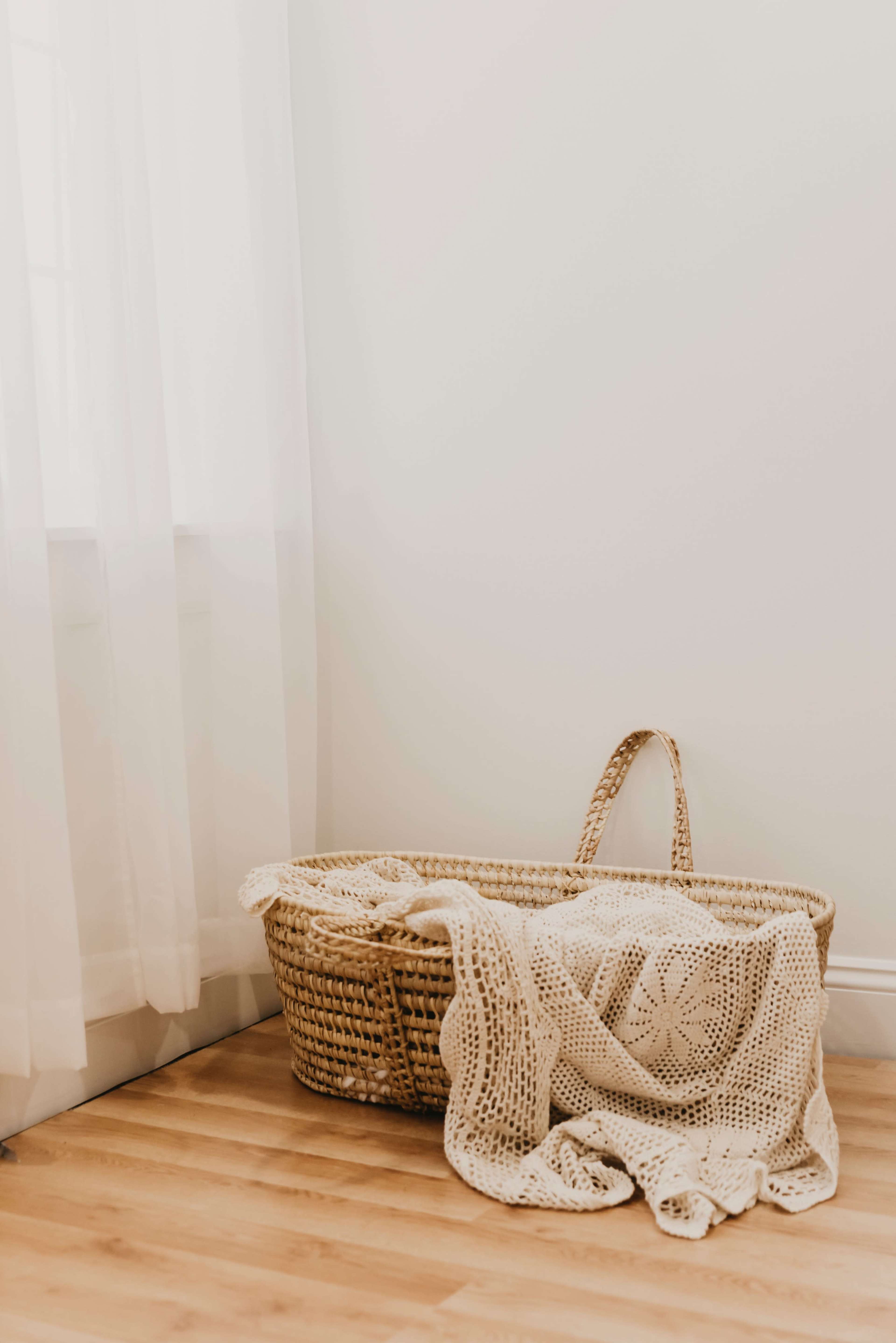 A woven basket sits on a wooden floor near a window, with a light-colored blanket draped over it.