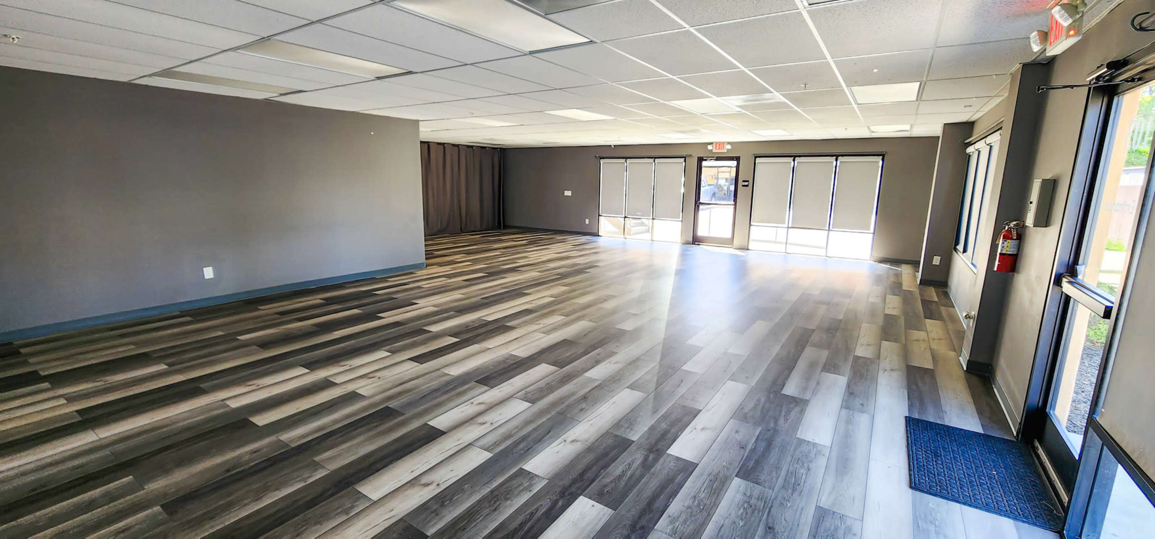 A spacious, empty room with a wood-like floor and large windows allowing natural light.