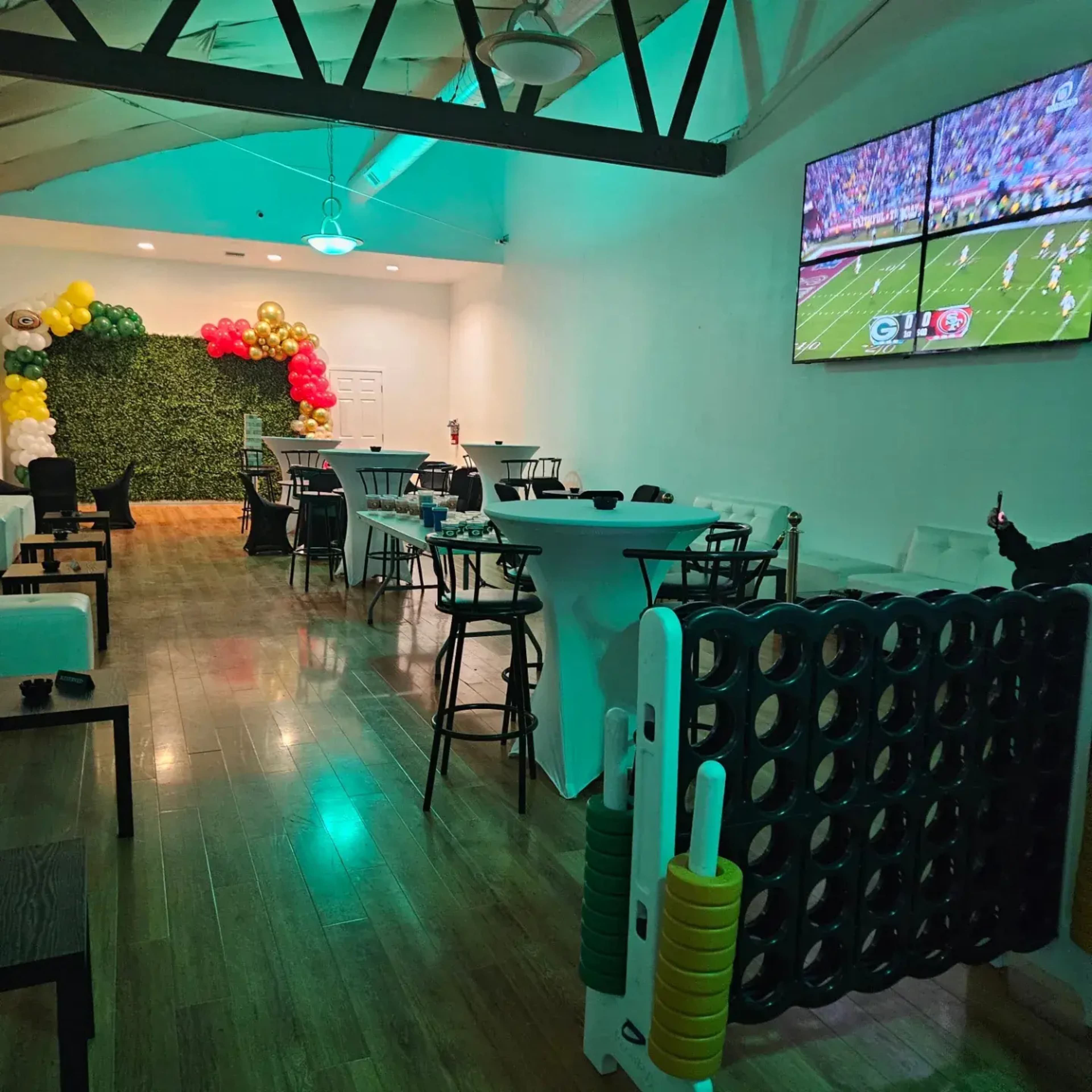 The image shows a spacious event room decorated with green foliage and colorful balloons, featuring multiple televisions displaying a sports game.