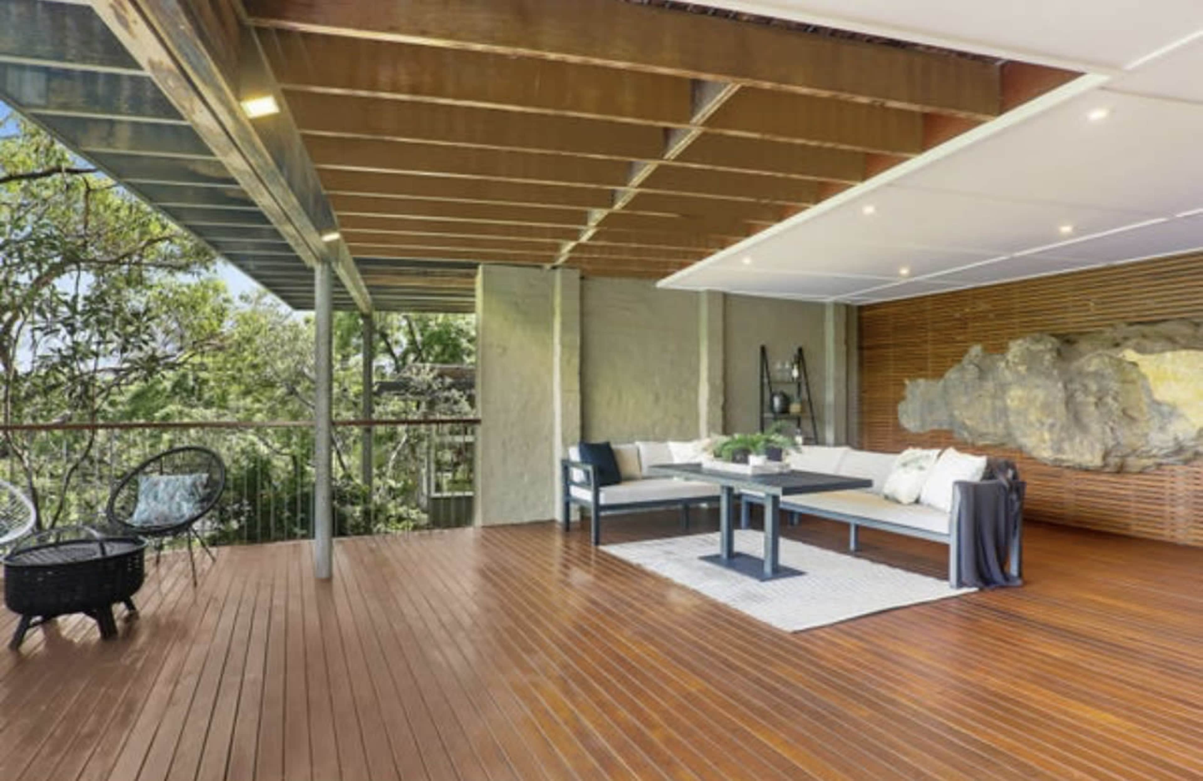 A spacious outdoor living area with wooden flooring, a modern seating arrangement, and greenery visible in the background.