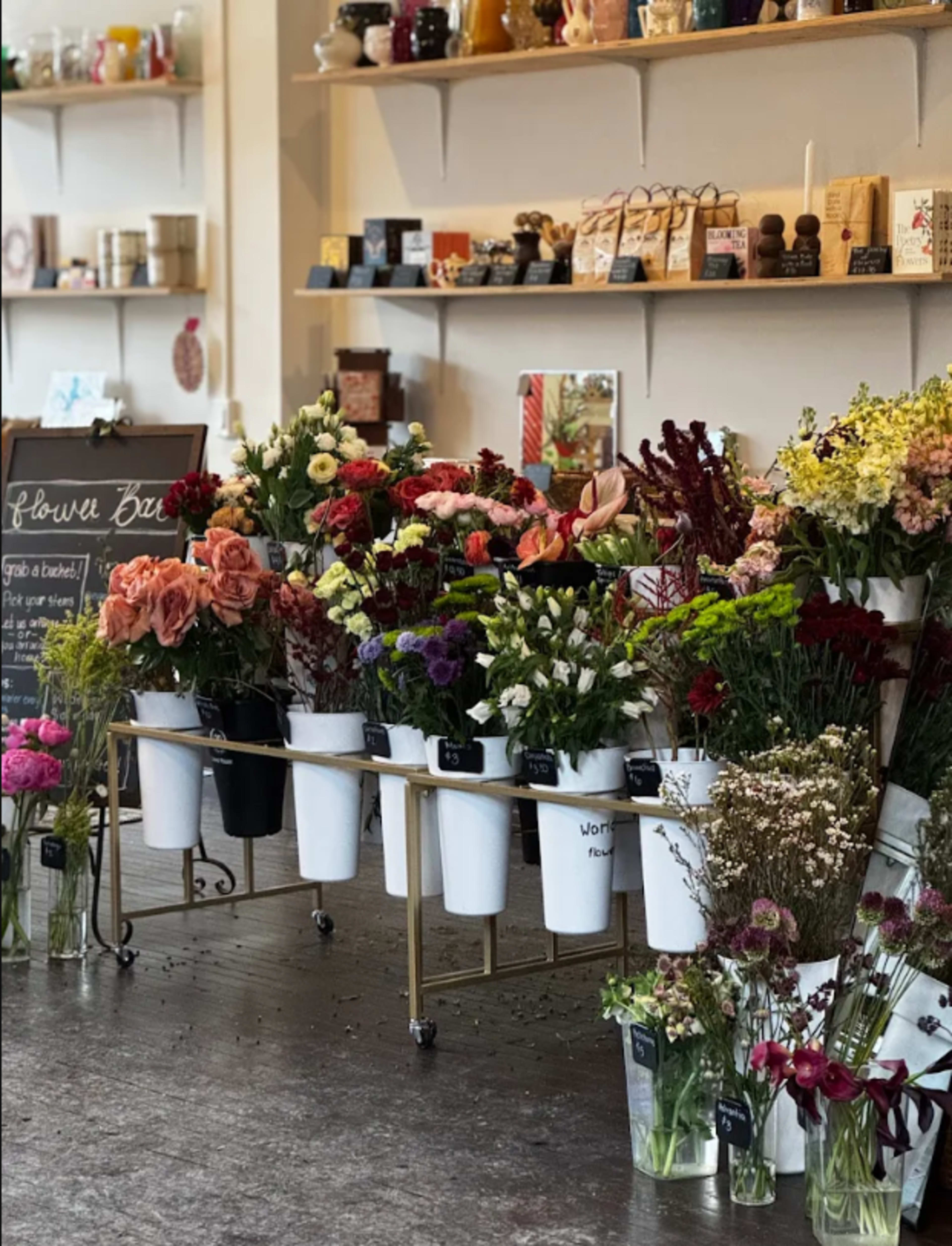 The image shows a selection of various flowers displayed in white pots on a cart, with shelves of decorative items and candles in the background.