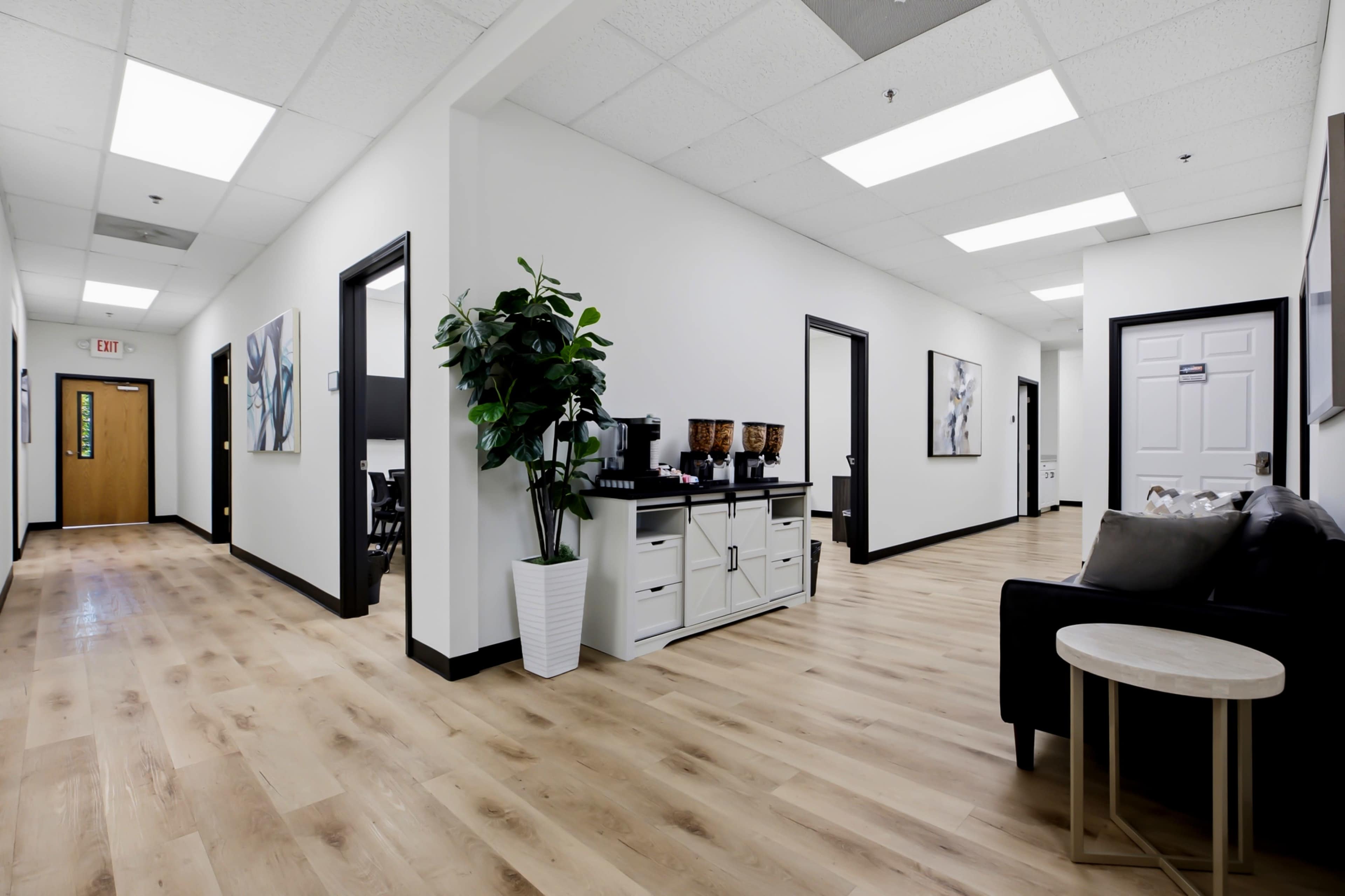 The image shows a brightly lit hallway in an office setting, featuring wooden floors, white walls, and a seating area with a plant and a small table.