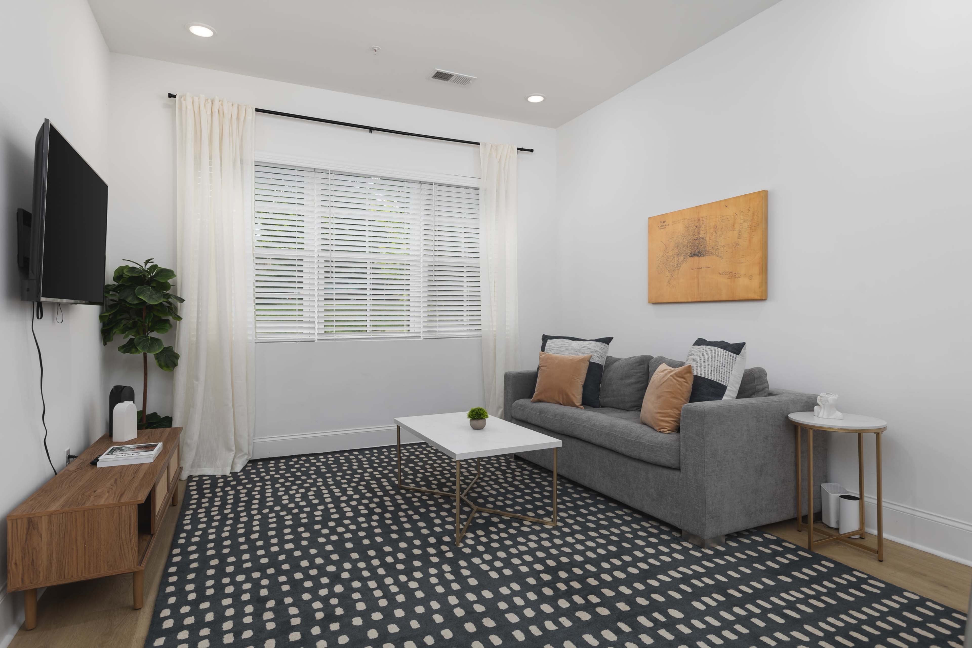 A modern living room features a gray sofa, a coffee table on a patterned rug, and a window with sheer curtains.