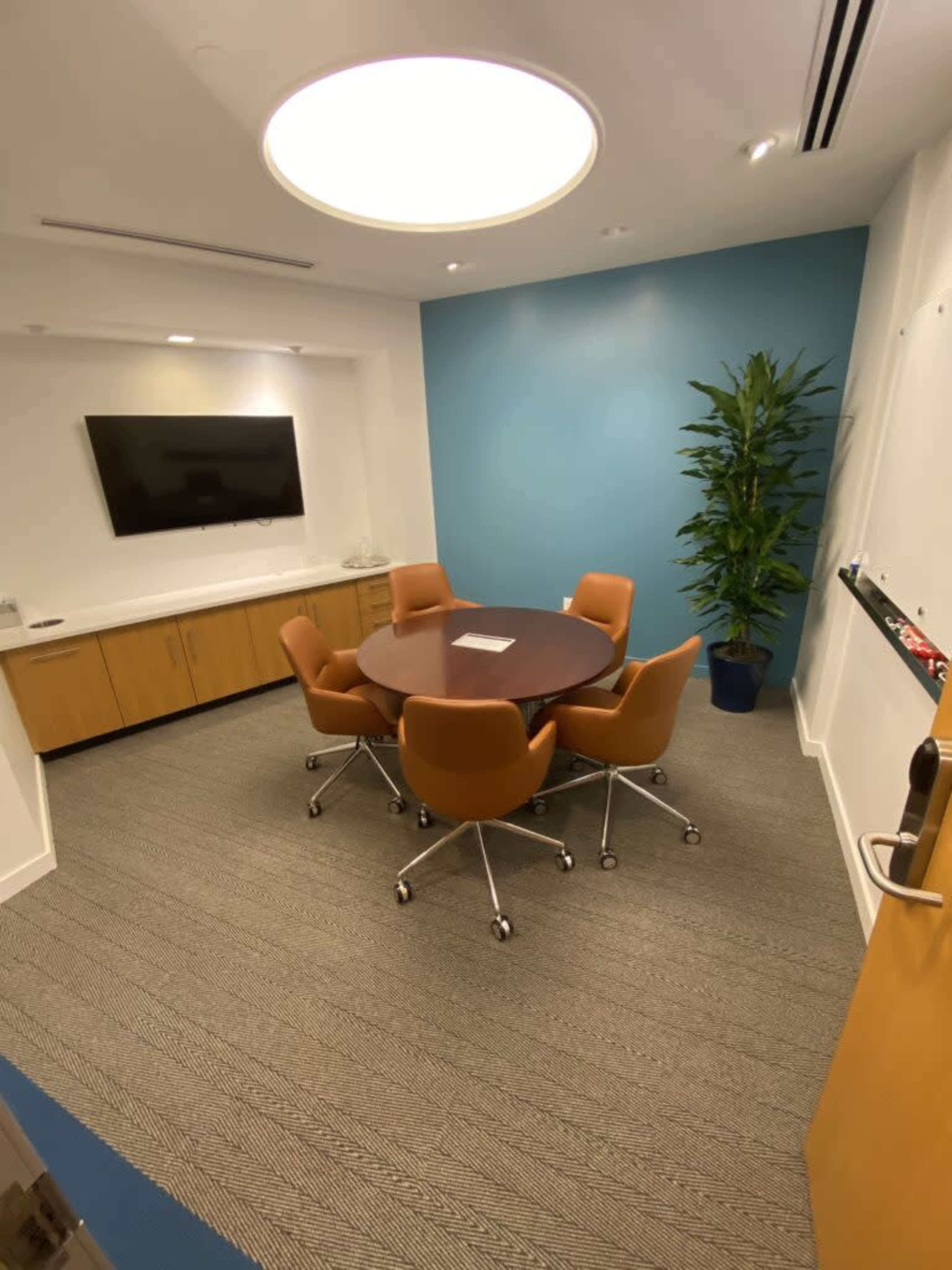 The image shows a small conference room with a round table surrounded by five orange chairs, a flat-screen TV on the wall, and a potted plant in the corner.