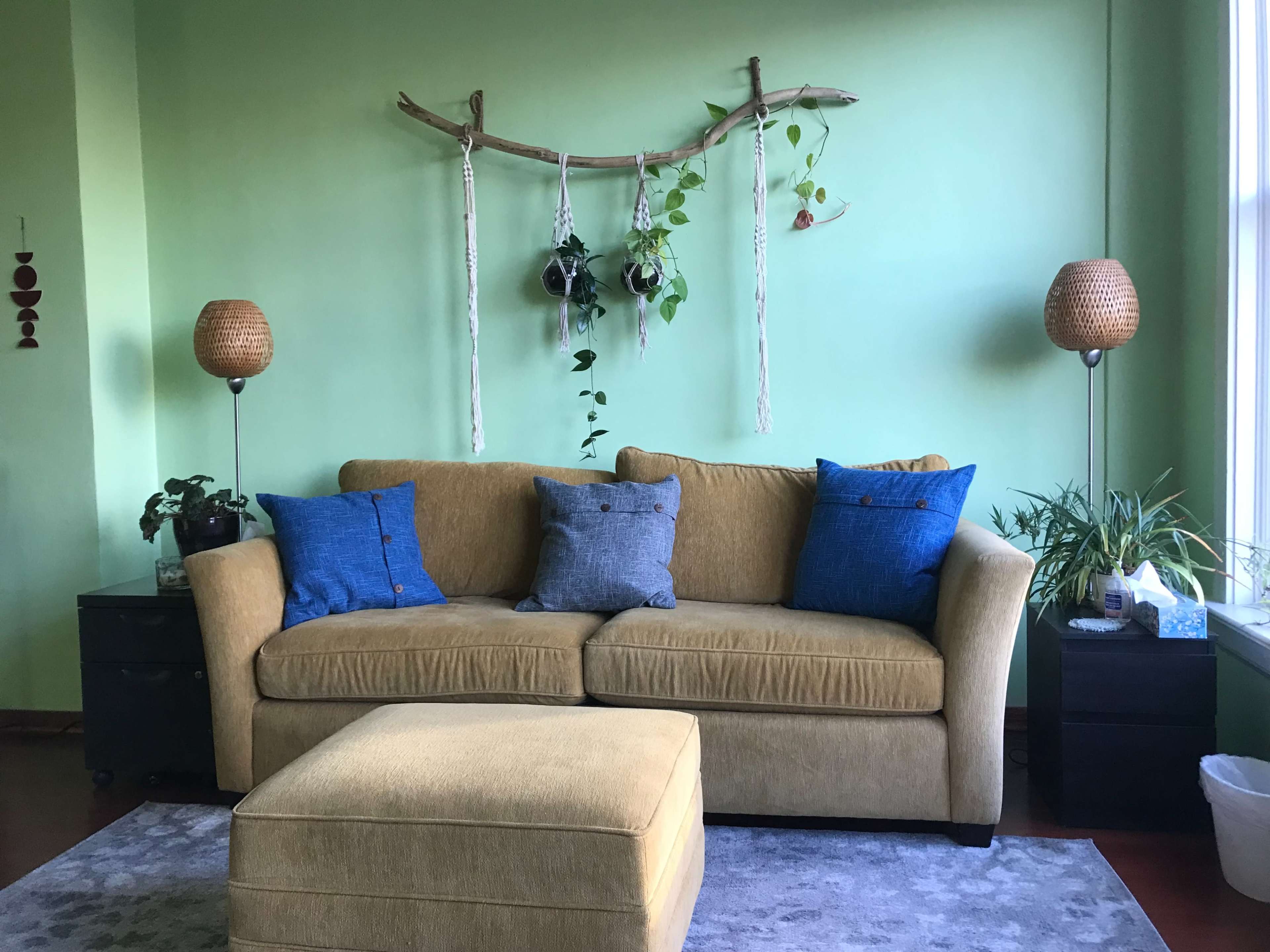 The image shows a living room with a green wall, a beige couch with blue and gray pillows, two wicker lamps, and a decorative hanging plant arrangement.