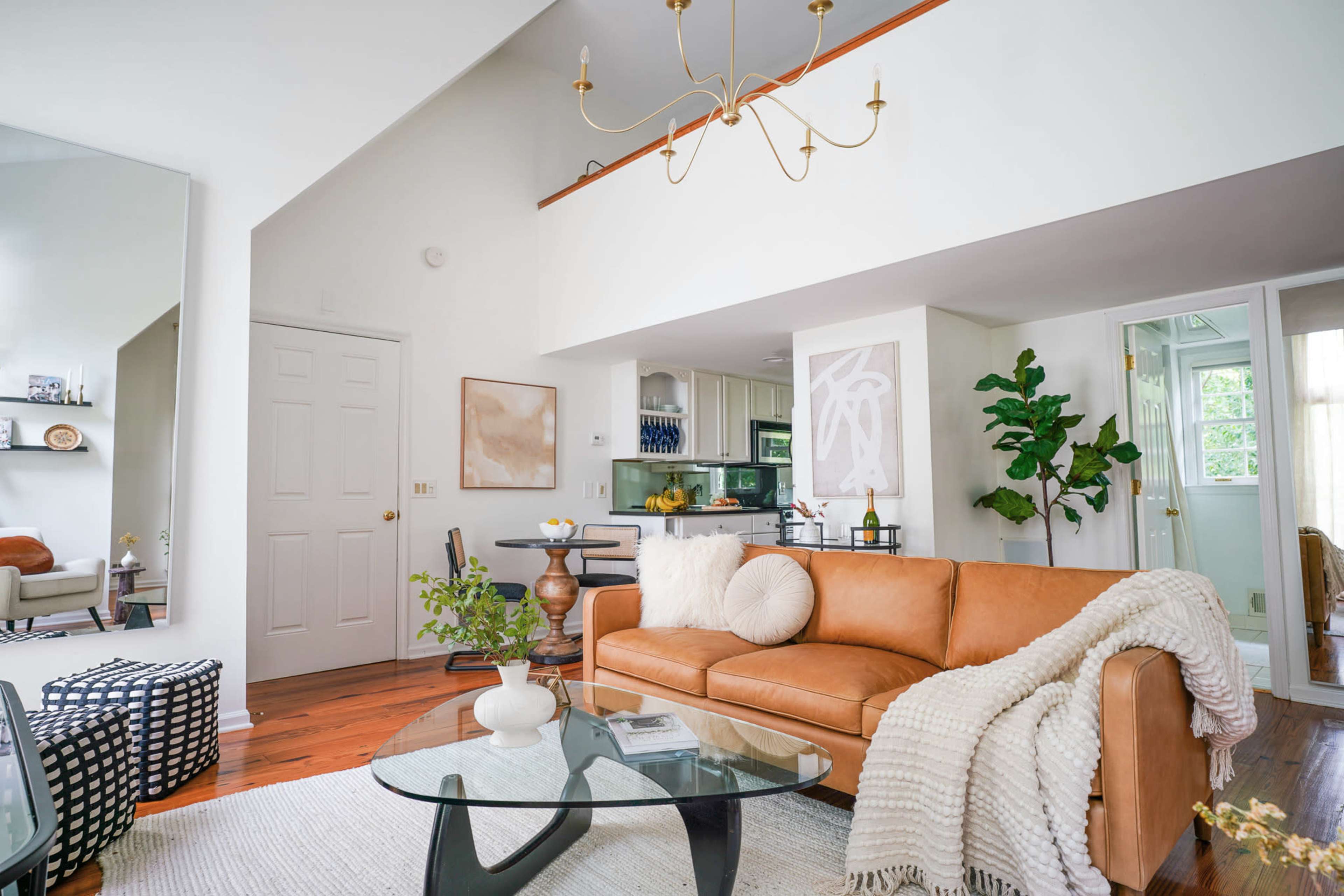A bright living room features a tan leather sofa, a glass coffee table, and a kitchen area visible in the background.