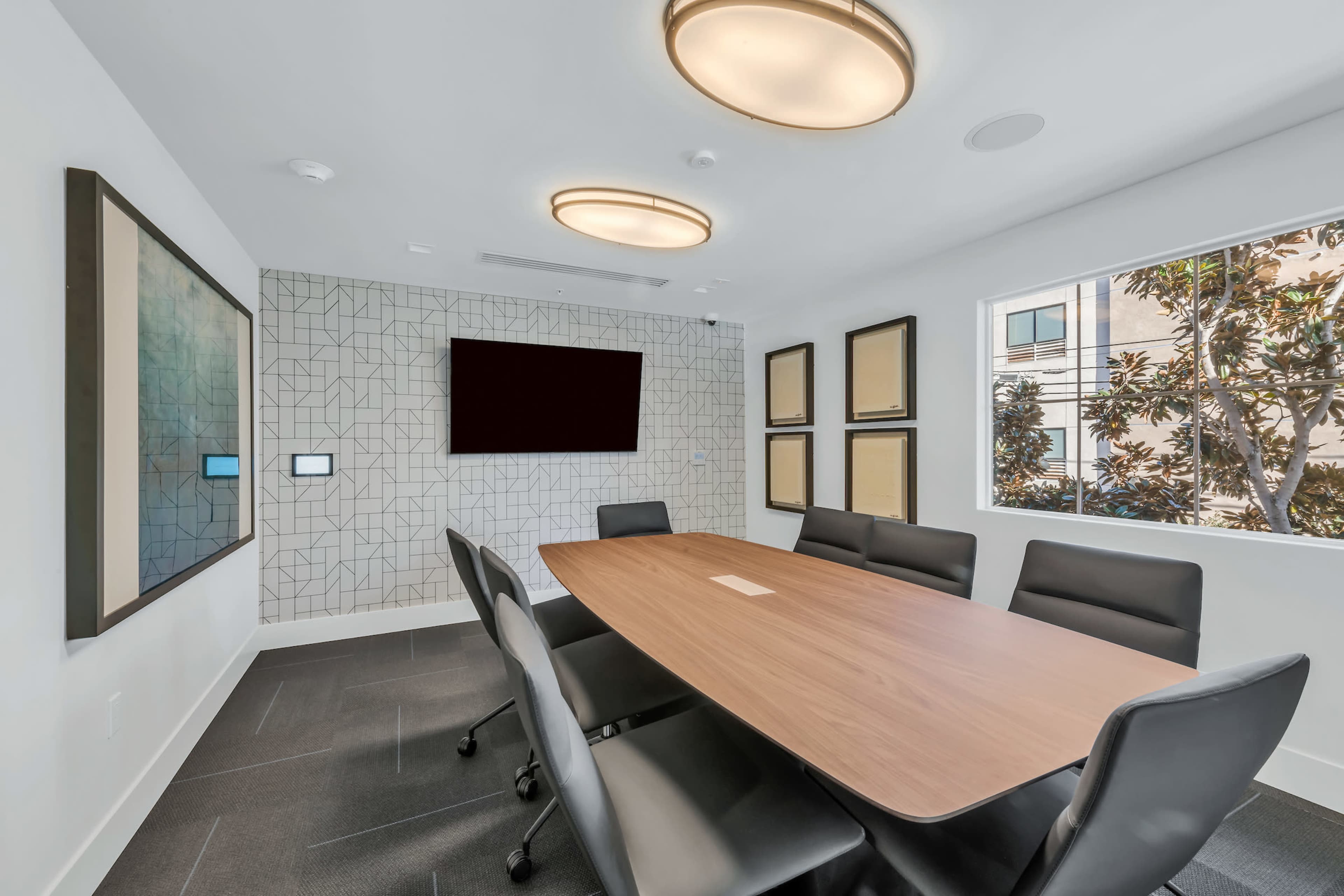 A modern conference room features a large wooden table surrounded by black chairs, with a wall-mounted screen and decorative framed panels.