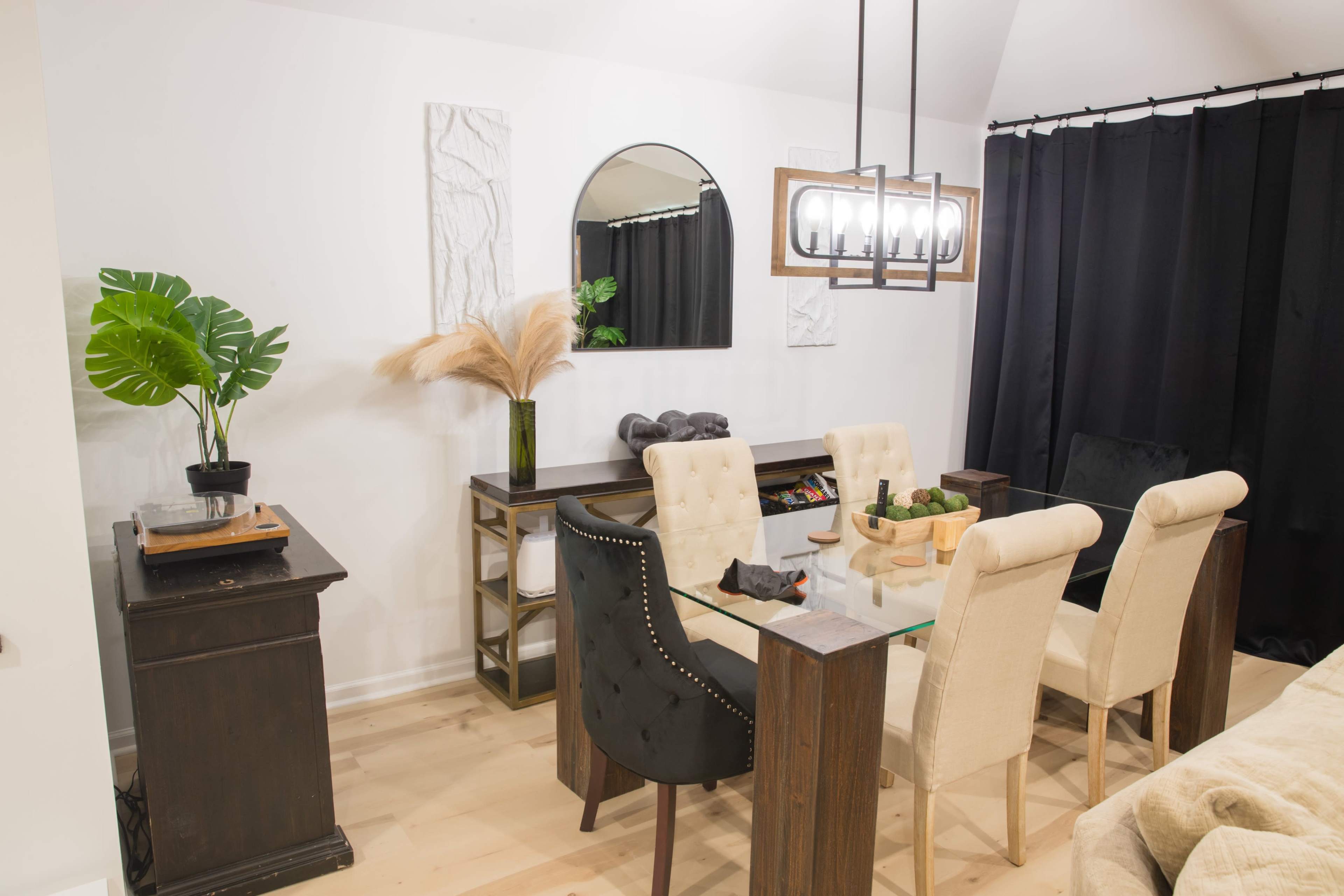 A modern dining area features a glass table surrounded by upholstered chairs, with a sideboard and decorative plants against a backdrop of black curtains.