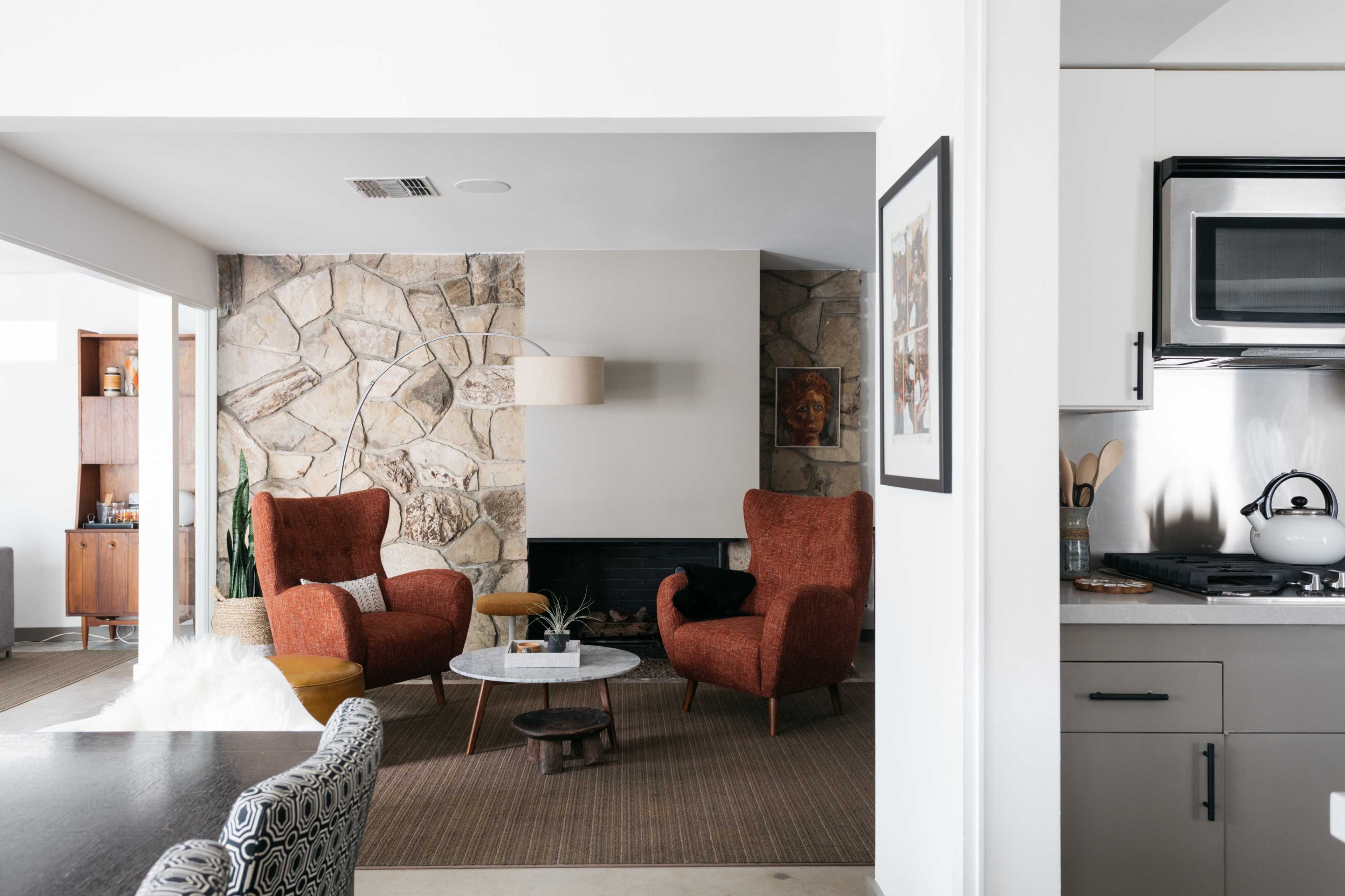 A modern living room features two orange armchairs facing a stone wall, with a coffee table and a kitchen area visible in the background.