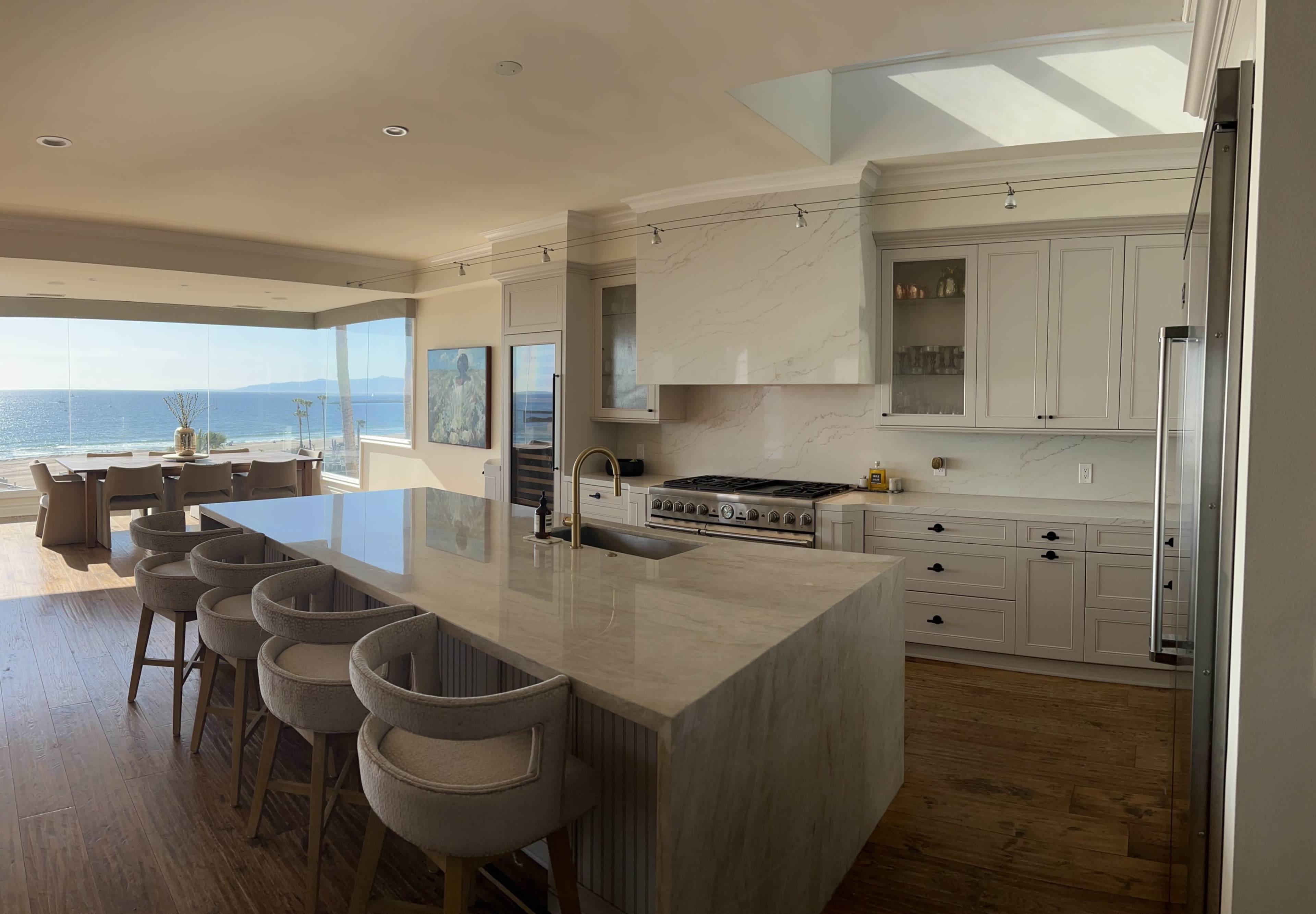A modern kitchen features a large marble island and panoramic windows overlooking the ocean.