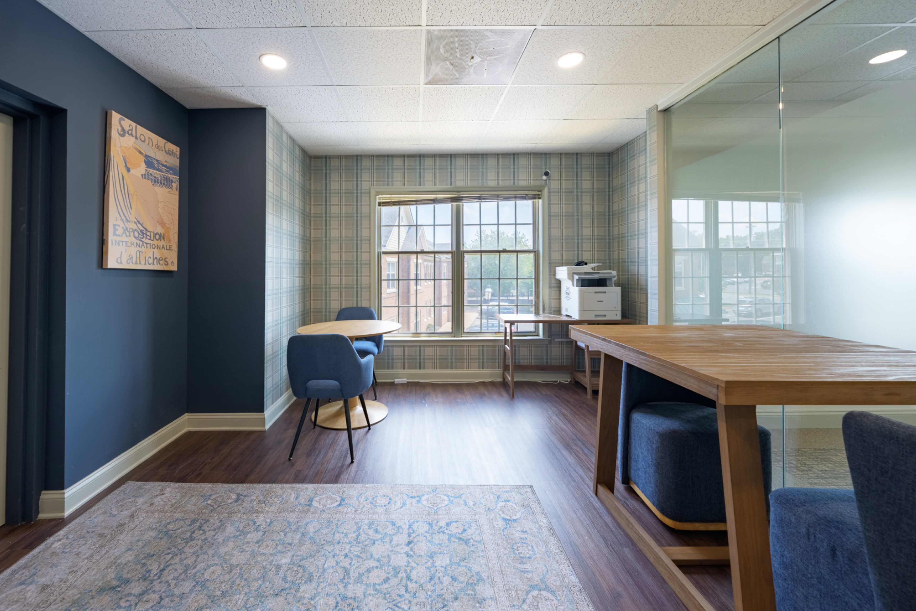 The image depicts a modern office space featuring a small seating area, a desk with a printer, and large windows allowing natural light to enter.