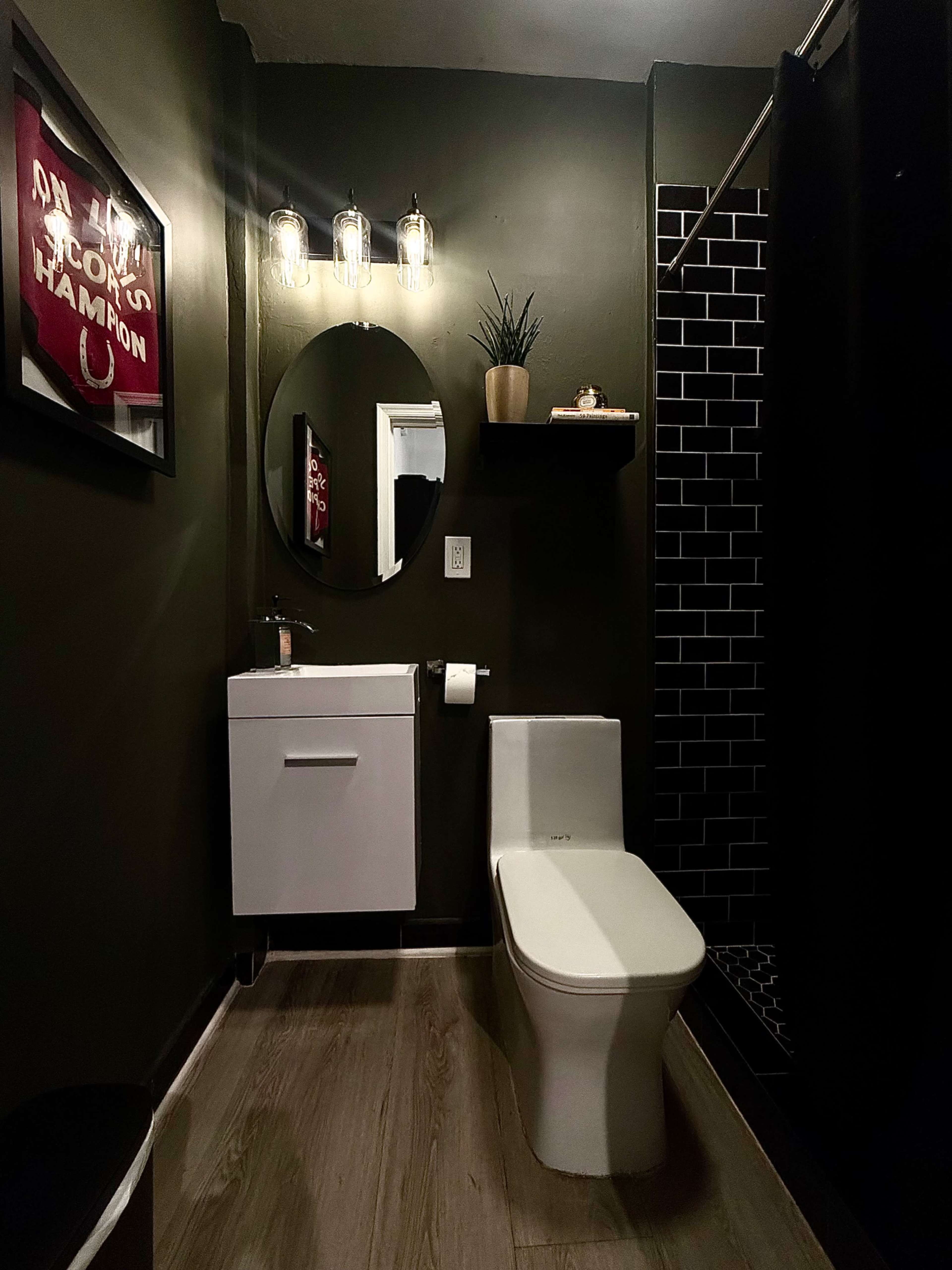 The image shows a modern bathroom featuring a black and white color scheme, a circular mirror, a wall-mounted vanity, and a standing shower with black tiles.