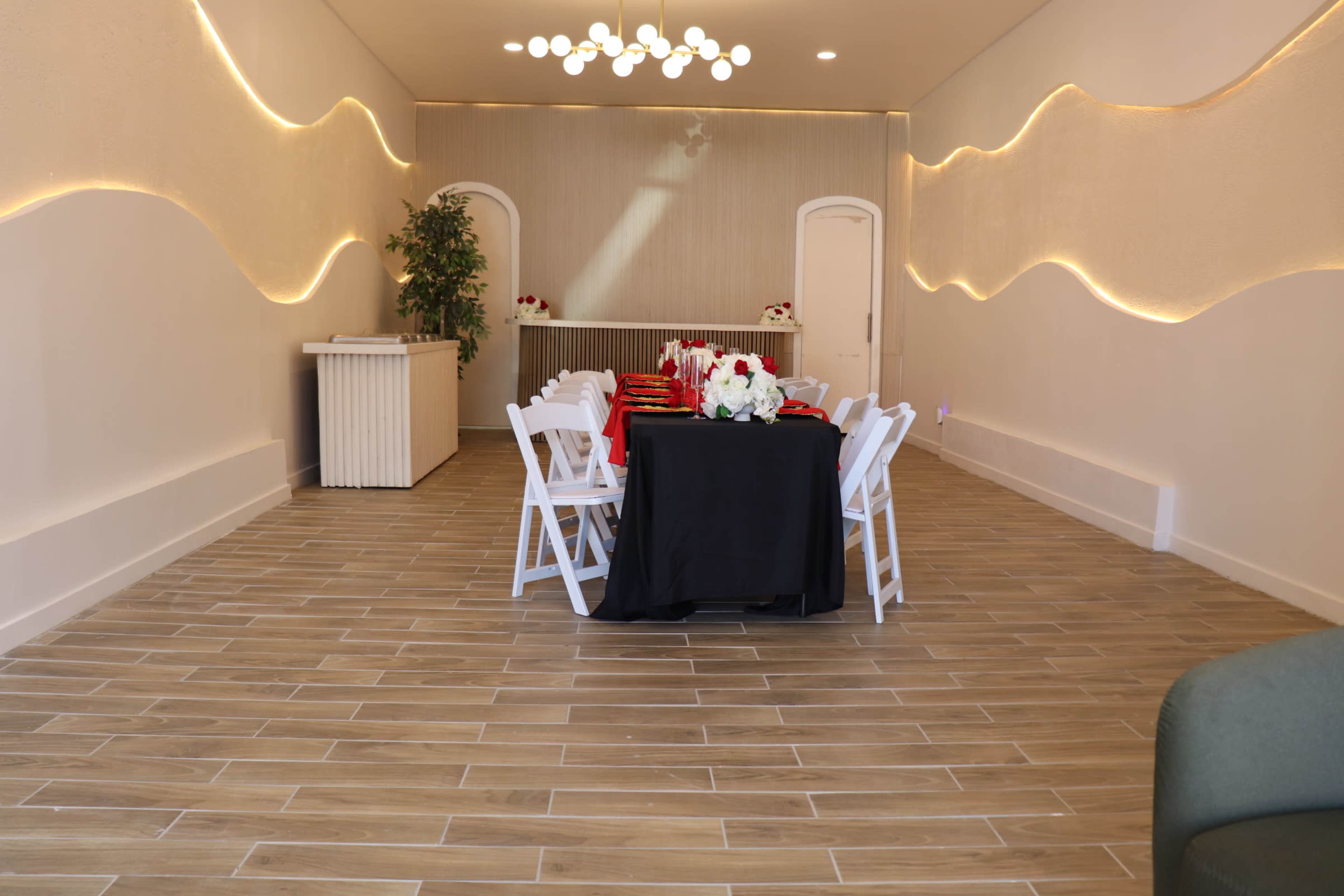 The image shows a well-lit interior room with a long table set for an event, surrounded by chairs, and decorative wall features.
