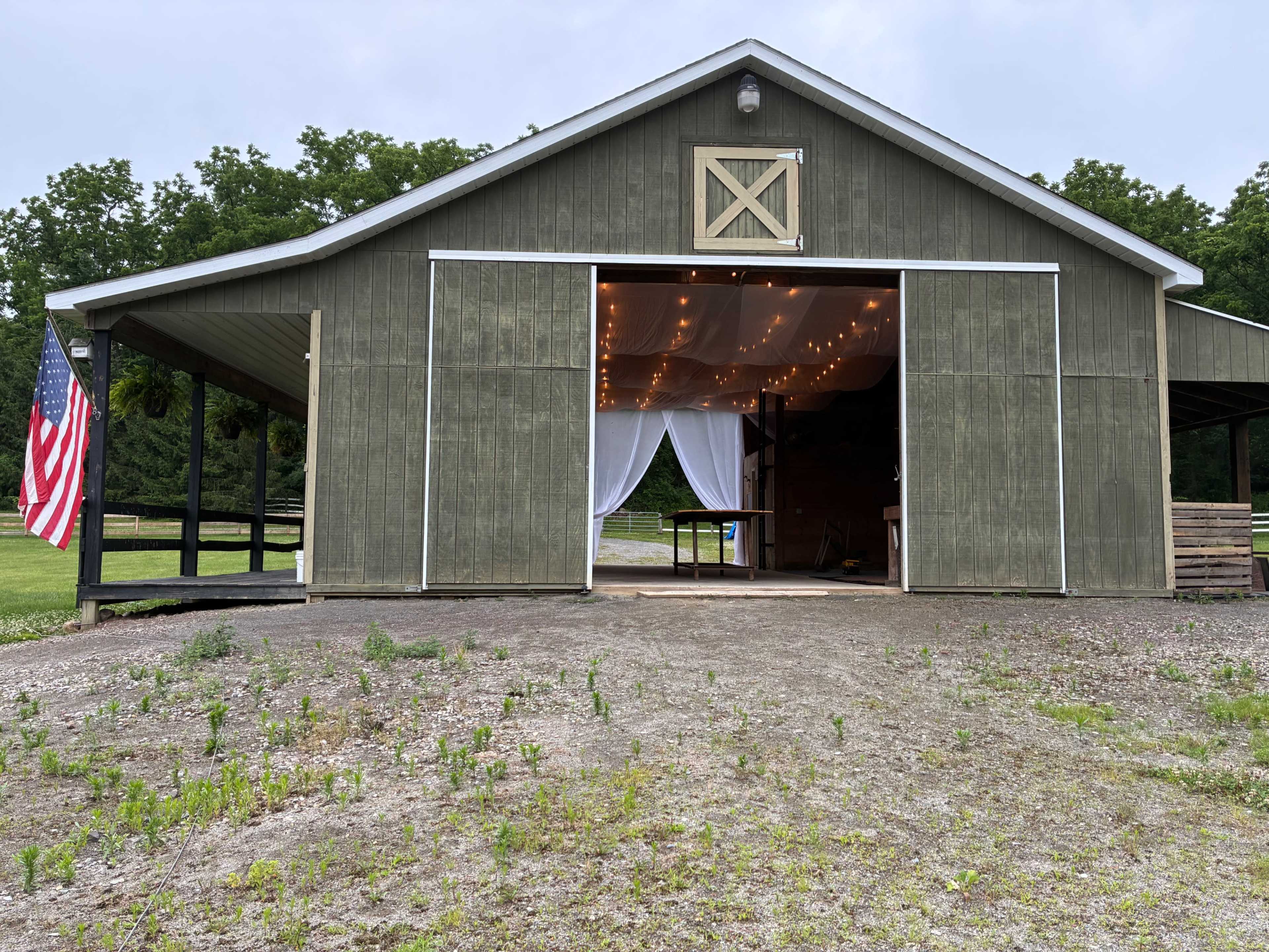 A large barn with an open front displays string lights hanging from the interior, set against a backdrop of greenery.
