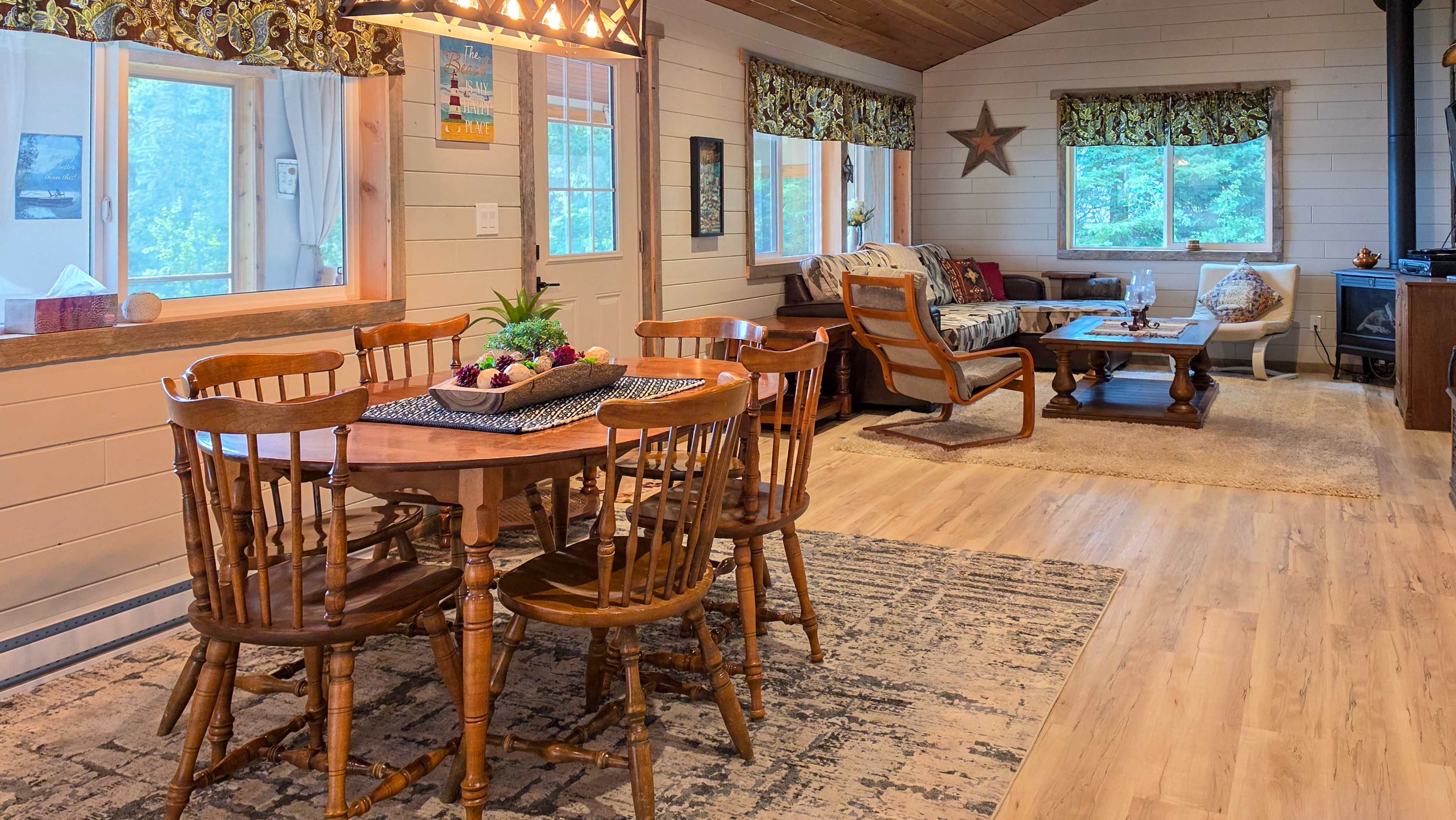 The image shows a cozy living space featuring a wooden dining table with chairs, a seating area with a couch and coffee table, and large windows allowing natural light to enter.
