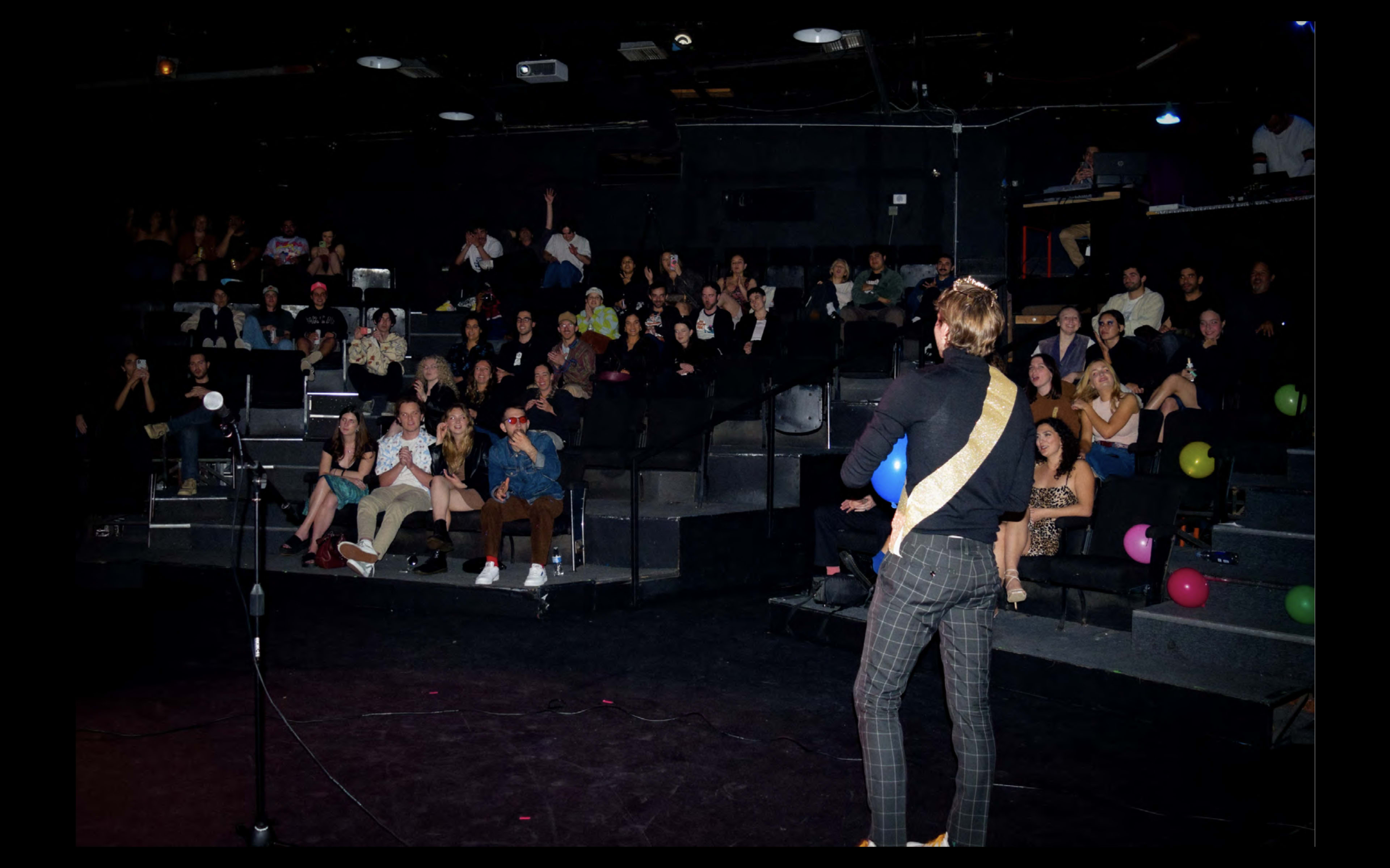 A performer with a sash stands on stage, facing an audience seated in a darkened theater filled with colorful balloons and illuminated by stage lights.