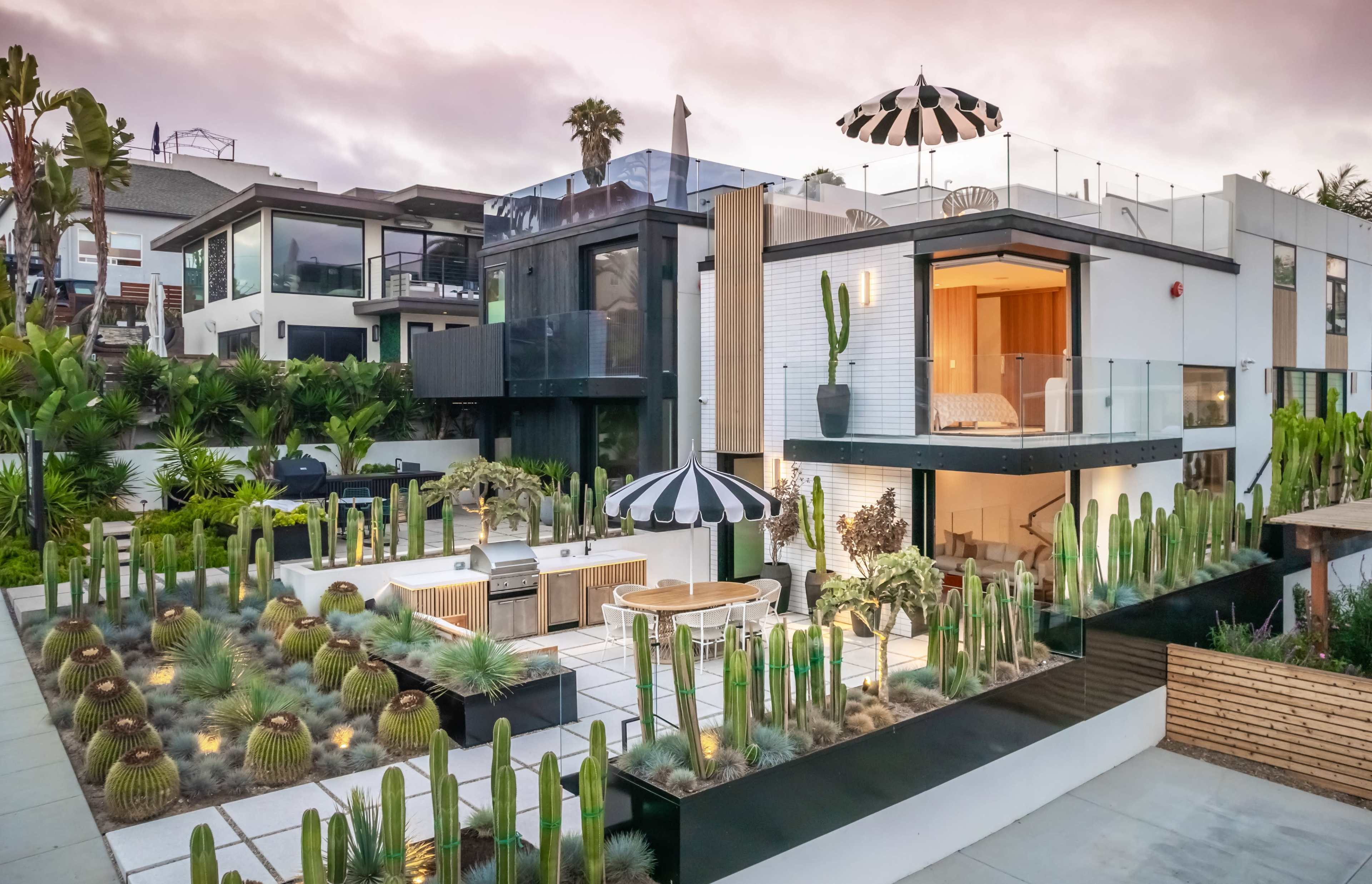 A modern two-story house with large windows is surrounded by a landscaped area featuring various cacti and a barbecue setup, topped with a black and white striped umbrella.