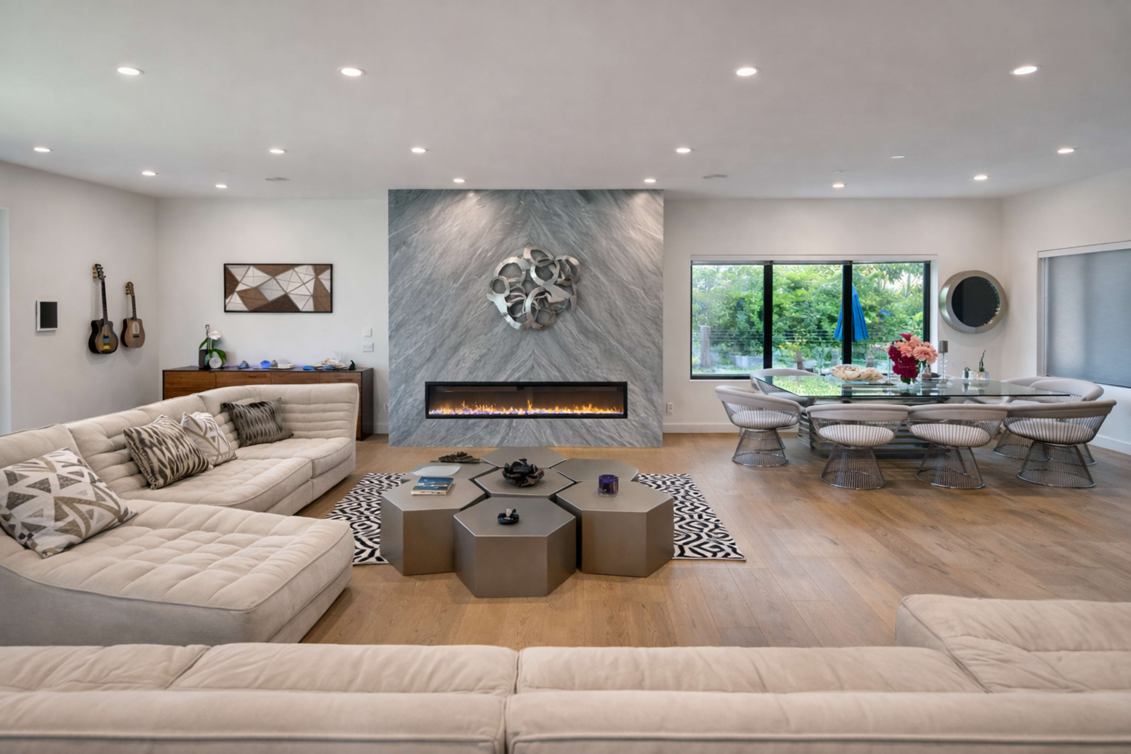 The image depicts a modern living room with a large sectional sofa, a geometric coffee table, an electric fireplace, and a dining area featuring a round table surrounded by chairs, all situated near a window overlooking greenery.