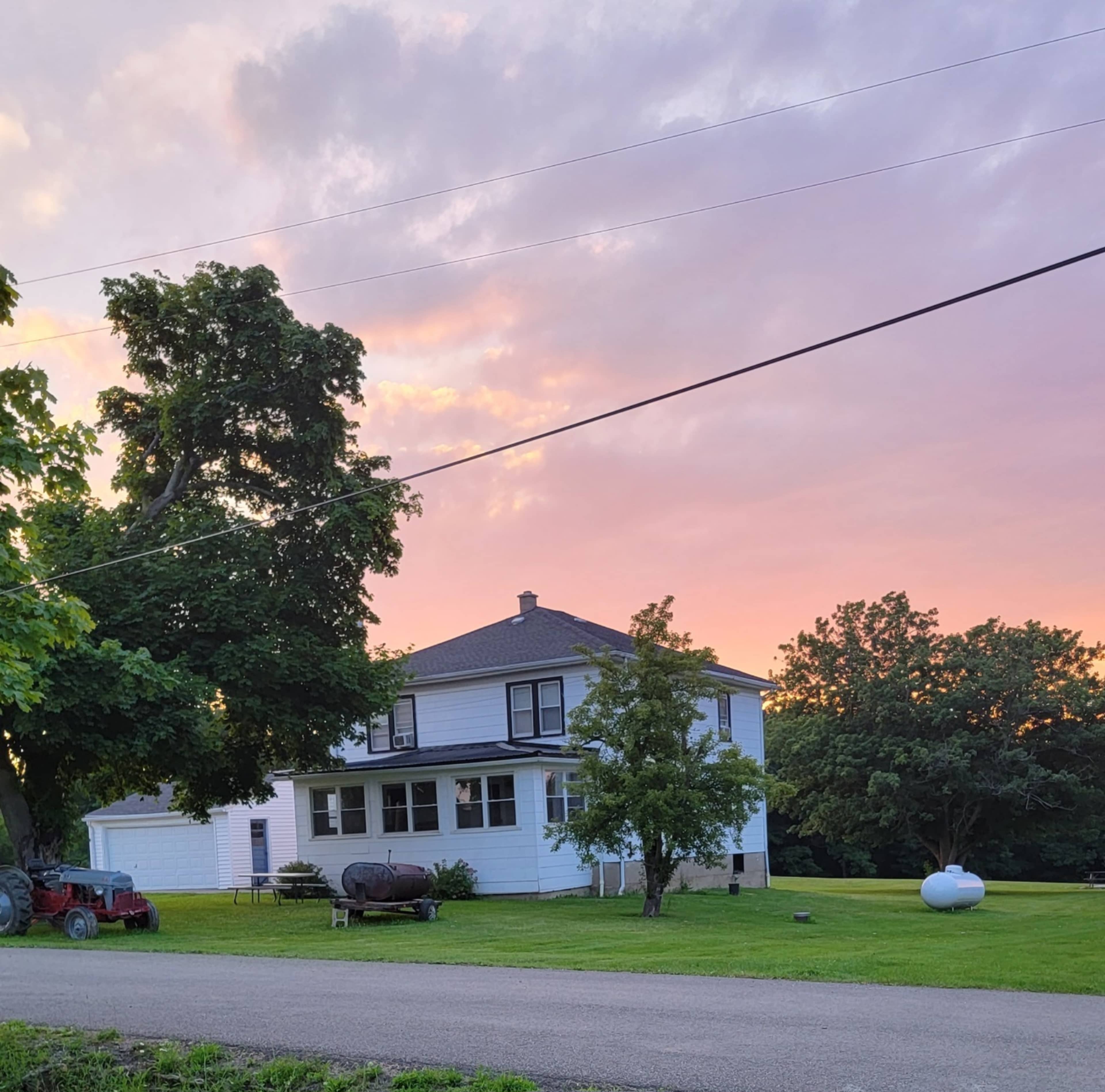 A two-story white farmhouse with a green lawn and nearby trees sits under a colorful sky at sunset.