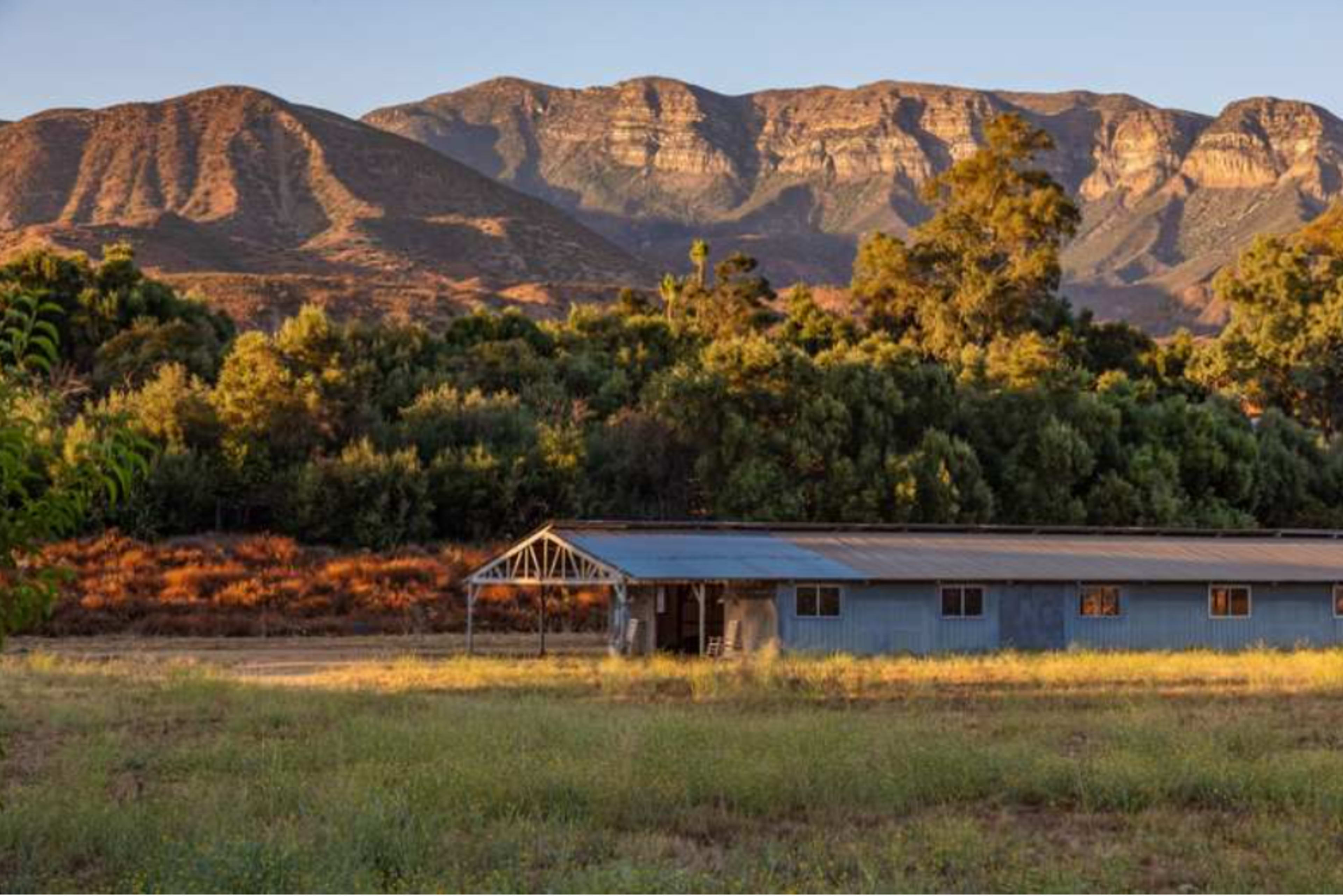 A weathered barn sits in a grassy field, with rugged mountains rising in the background.