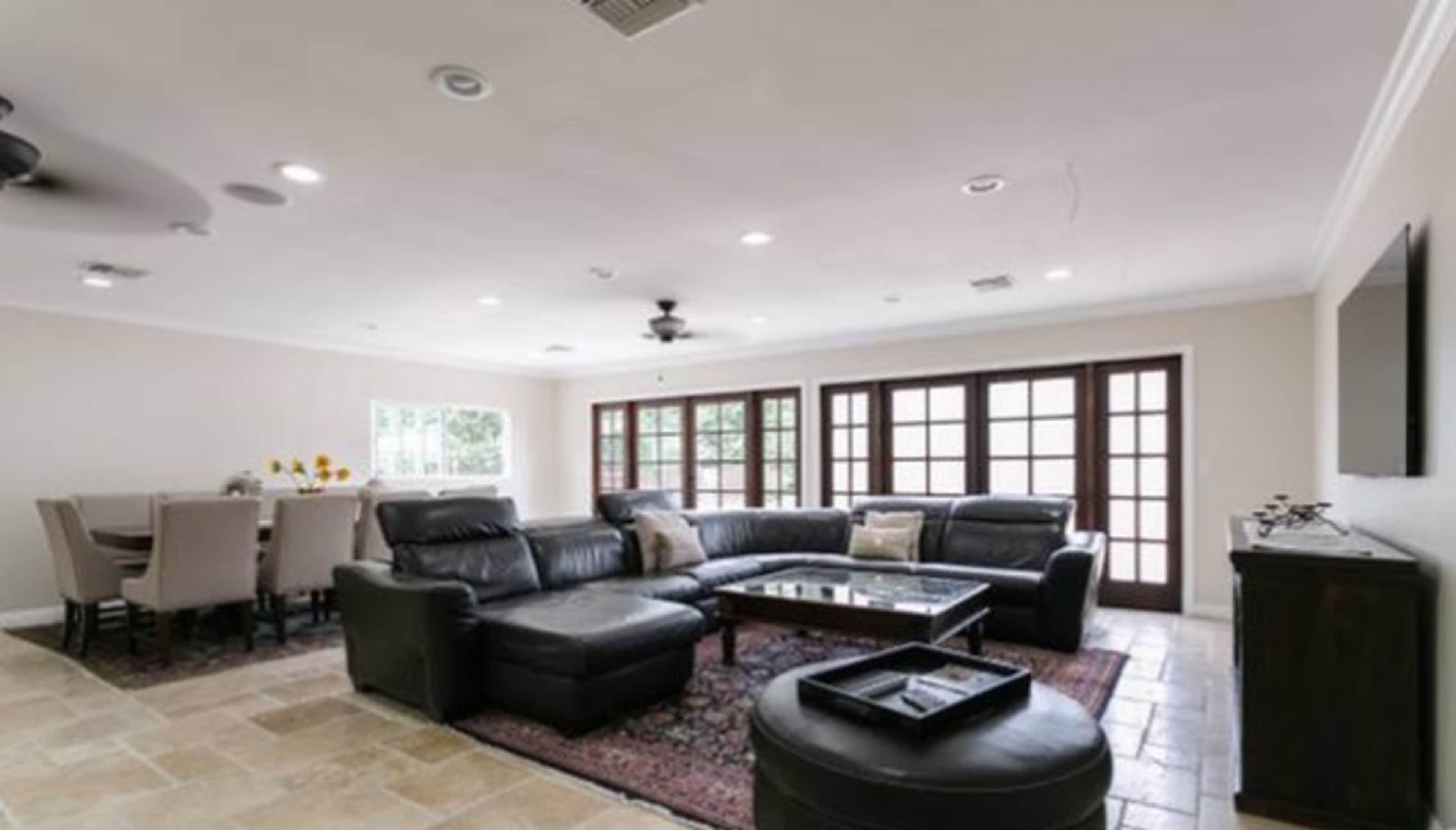 The image shows a living room with a sectional leather couch, a coffee table, and a dining area with a table and chairs, illuminated by natural light from multiple windows.