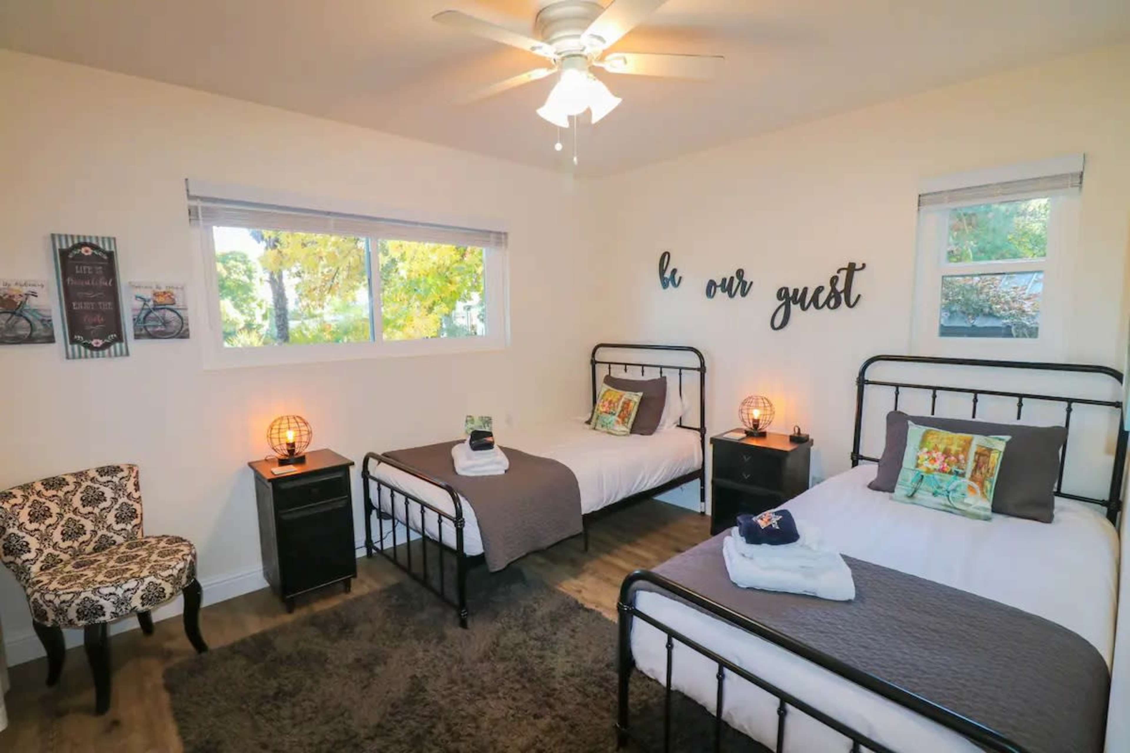 The room features two twin beds with dark bedding, a chair, a nightstand on each side, and a window that allows natural light to enter.