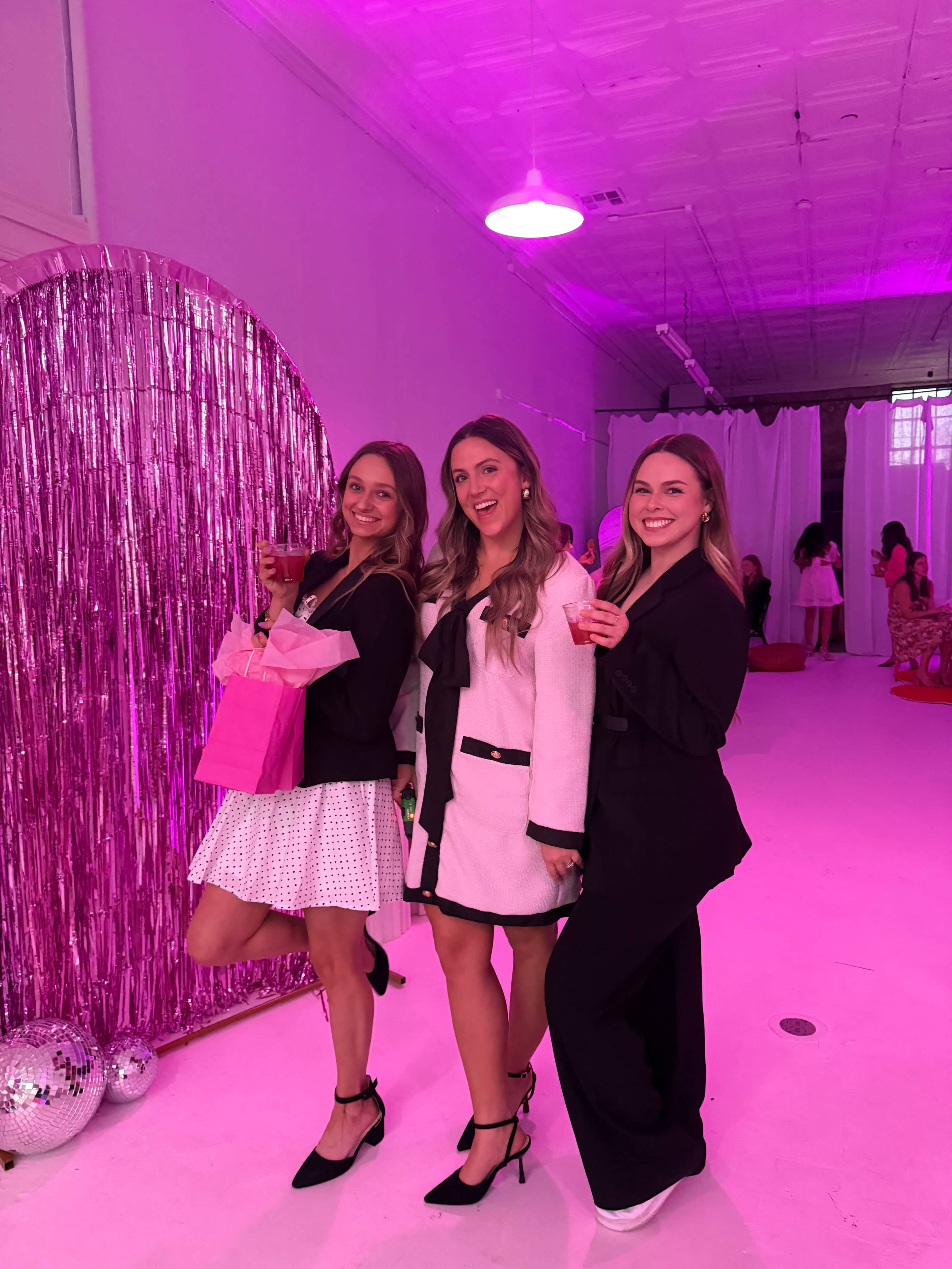 Three women pose together in a brightly lit room decorated with pink accents, holding drinks and a gift bag.