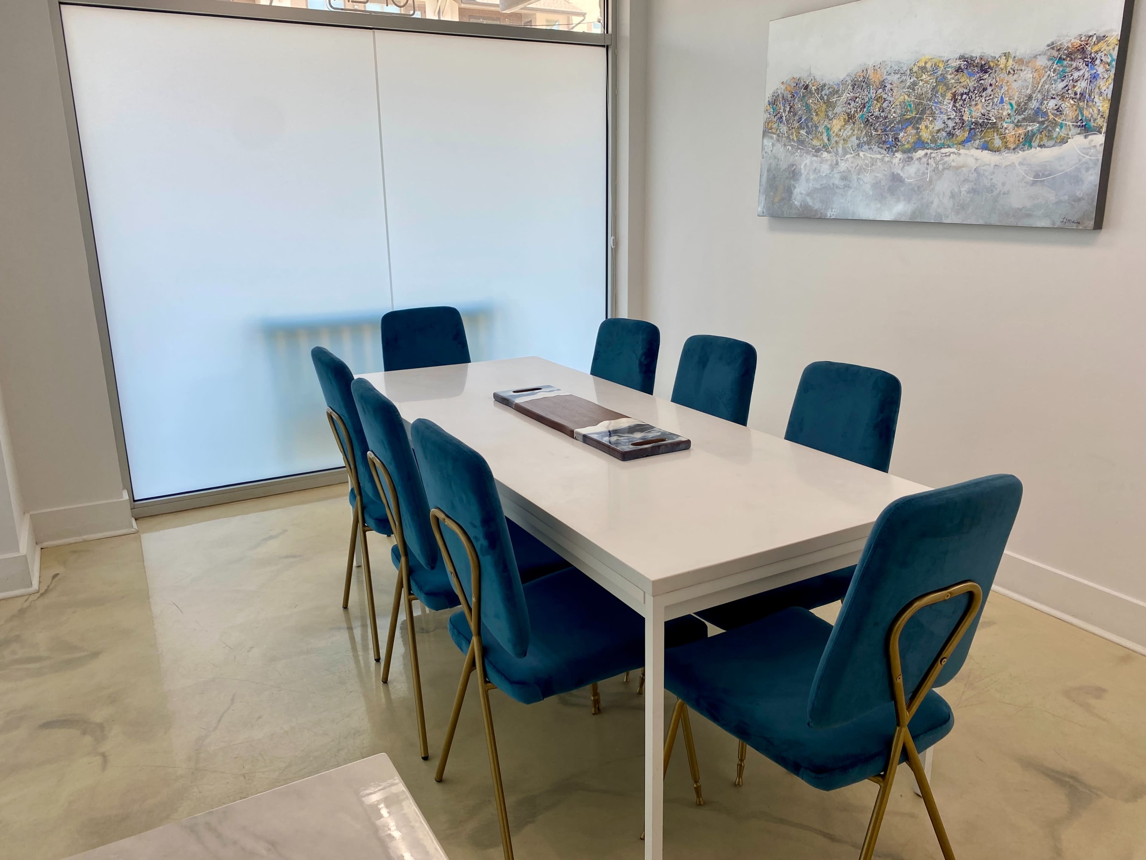 A modern dining room features a white table surrounded by eight teal chairs, with a large piece of abstract art on the wall and a sliding glass door in the background.