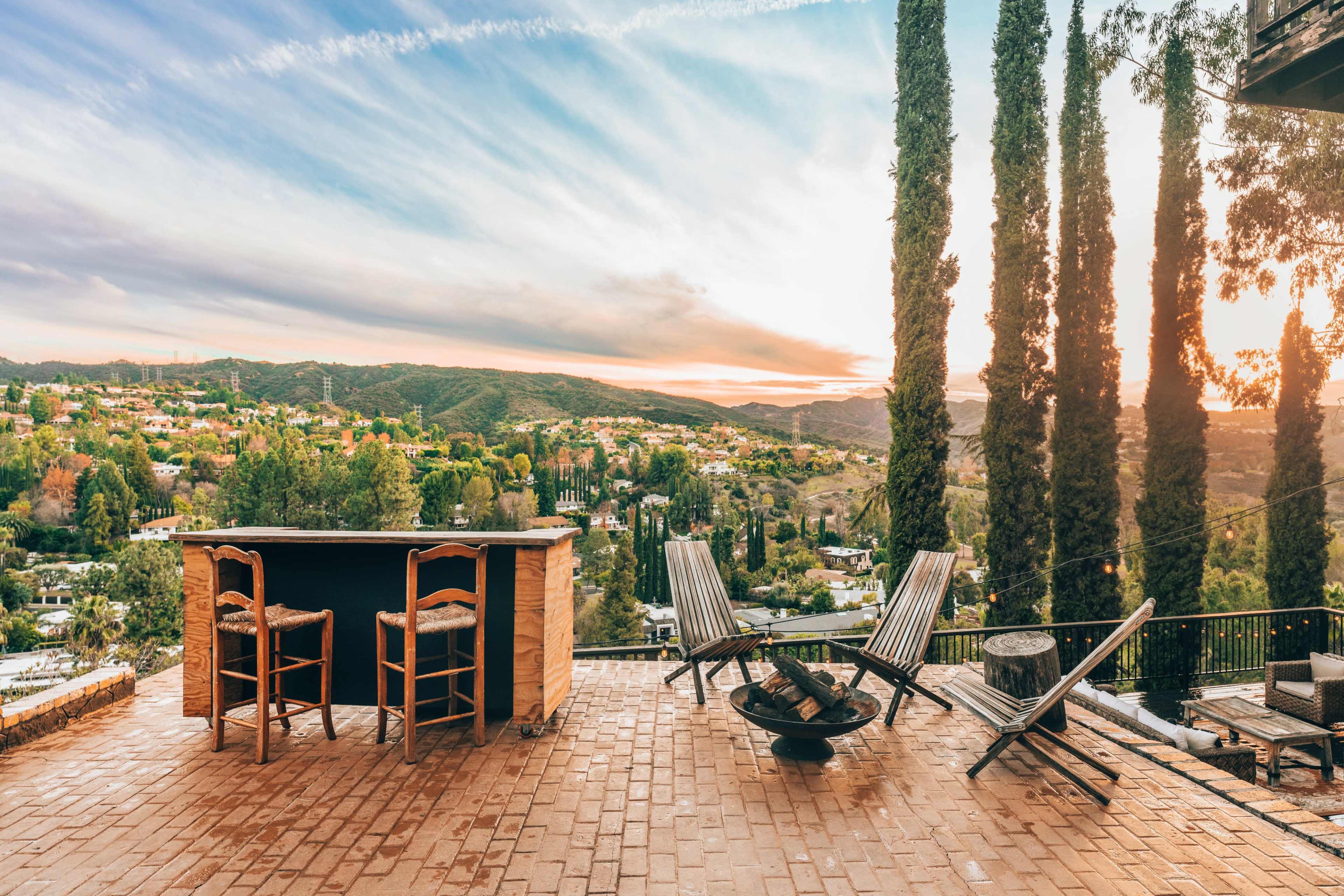 A patio with wooden chairs overlooks a mountainous landscape at sunset, framed by tall cypress trees.