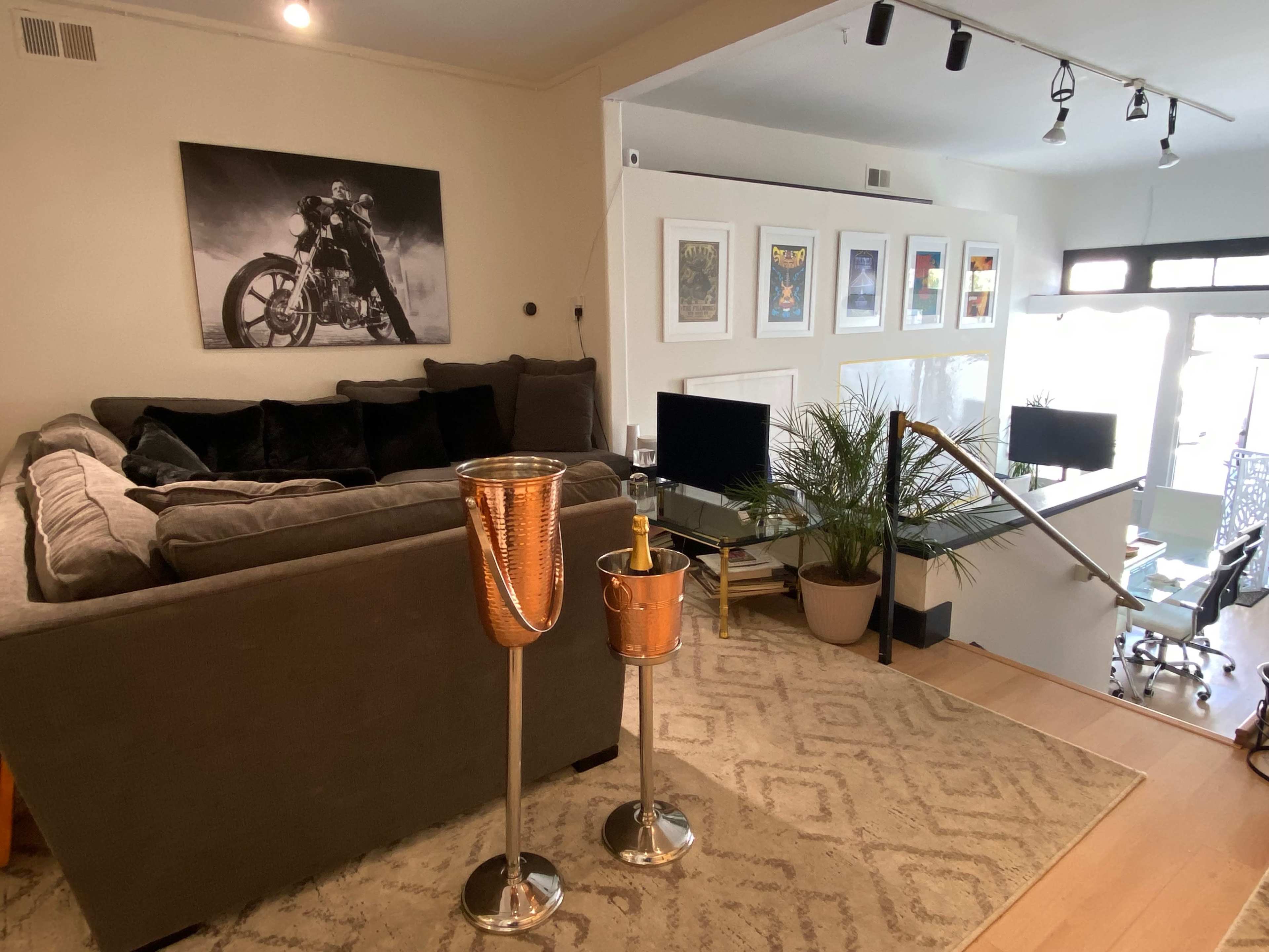 The image shows a cozy living area with a large gray sofa, a wall of framed art, two decorative stands with champagne buckets, and a glass railing leading to a bright workspace.