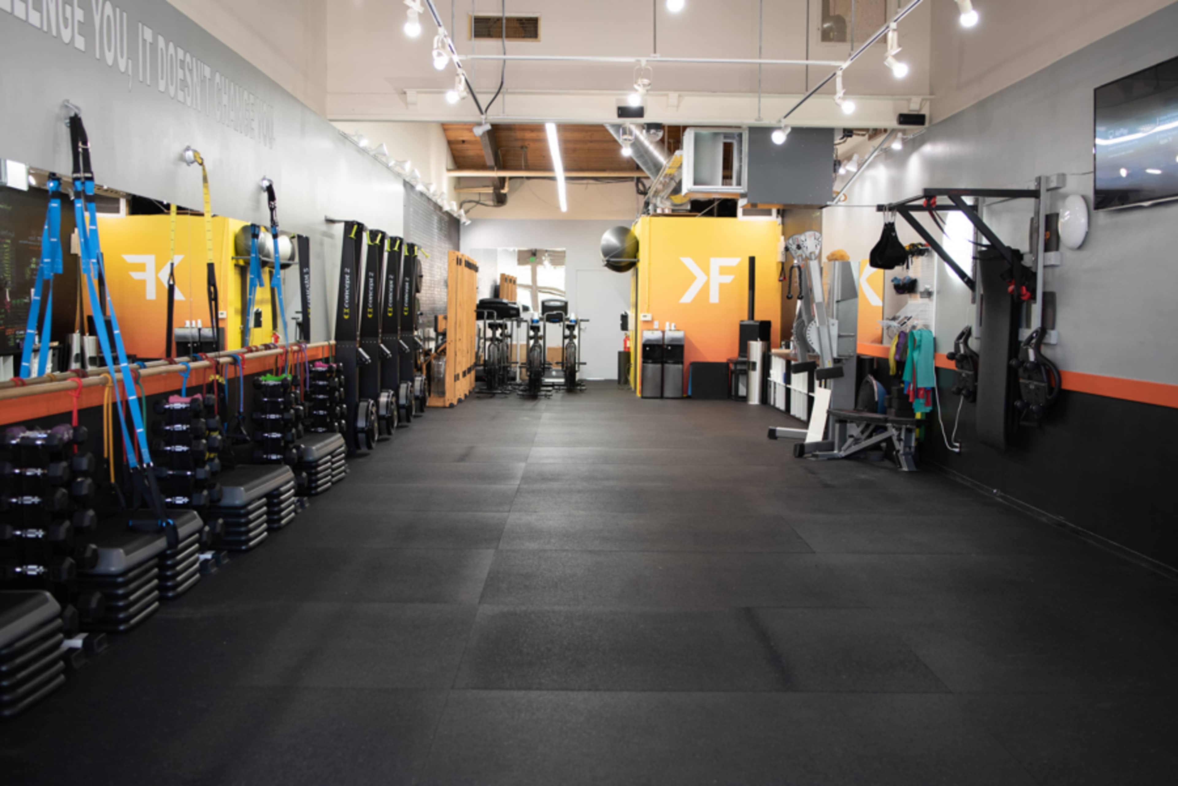 The image shows a gym interior with various exercise equipment organized along the walls and an open central area.